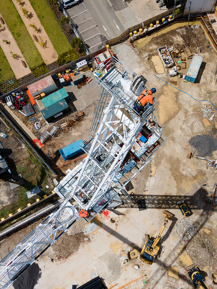 Aerial view of construction site with two workers in orange safety gear inspecting a white tower crane, surrounded by machinery and materials.