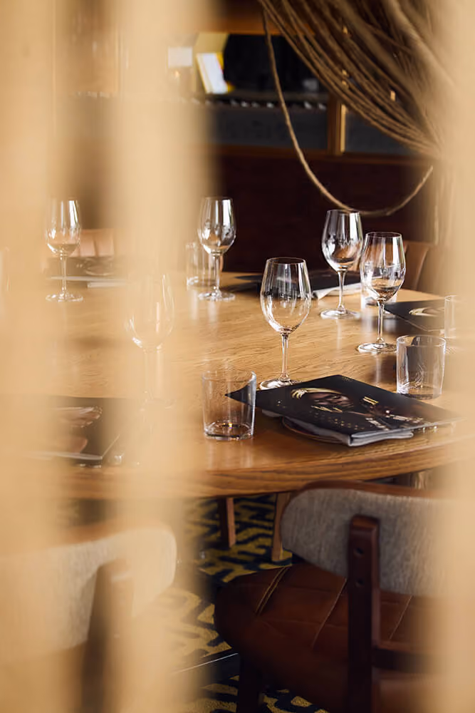 Close-up of a wooden dining table set with empty wine glasses, water glasses, menus, and cushioned chairs in a warmly lit restaurant.