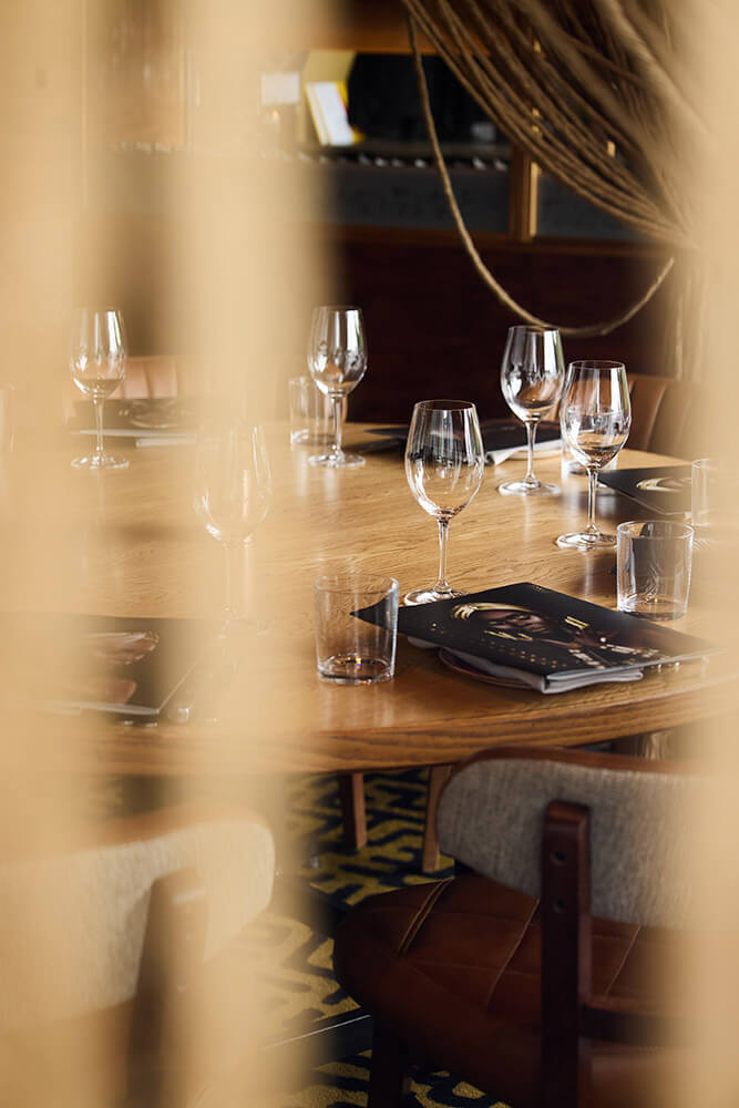 Close-up of a wooden dining table set with empty wine glasses, water glasses, menus, and cushioned chairs in a warmly lit restaurant.
