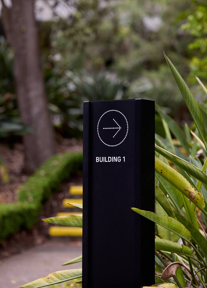 Black directional signpost with a right arrow and text 'BUILDING 1' surrounded by greenery and a pathway.