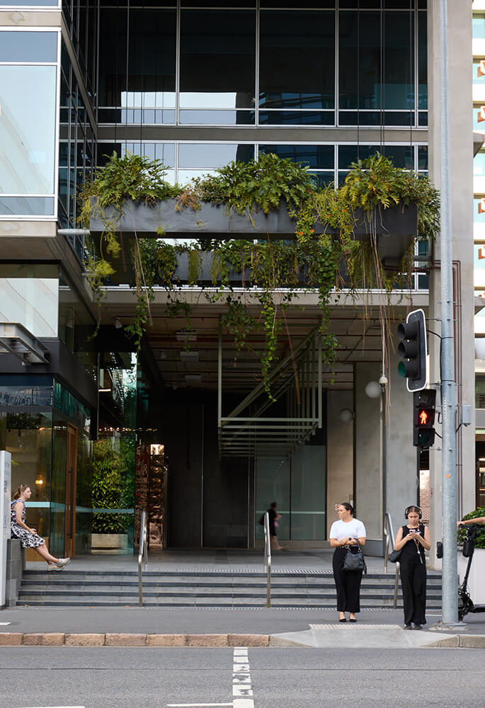 Modern building entrance with hanging plants above, and three people near steps and crosswalk, one sitting and two standing.