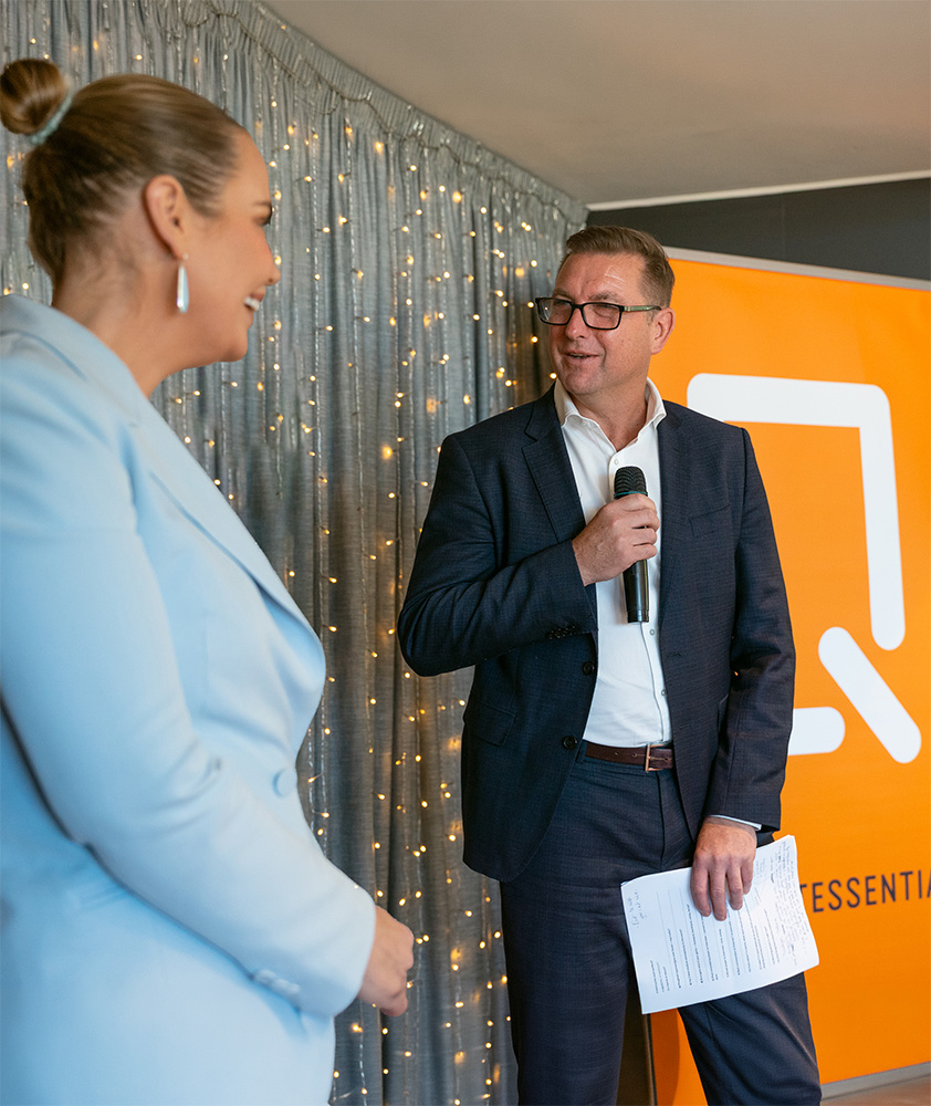 Man in a dark suit holding a microphone and papers speaking to a smiling woman in a light blue blazer against a backdrop of string lights and an orange banner.