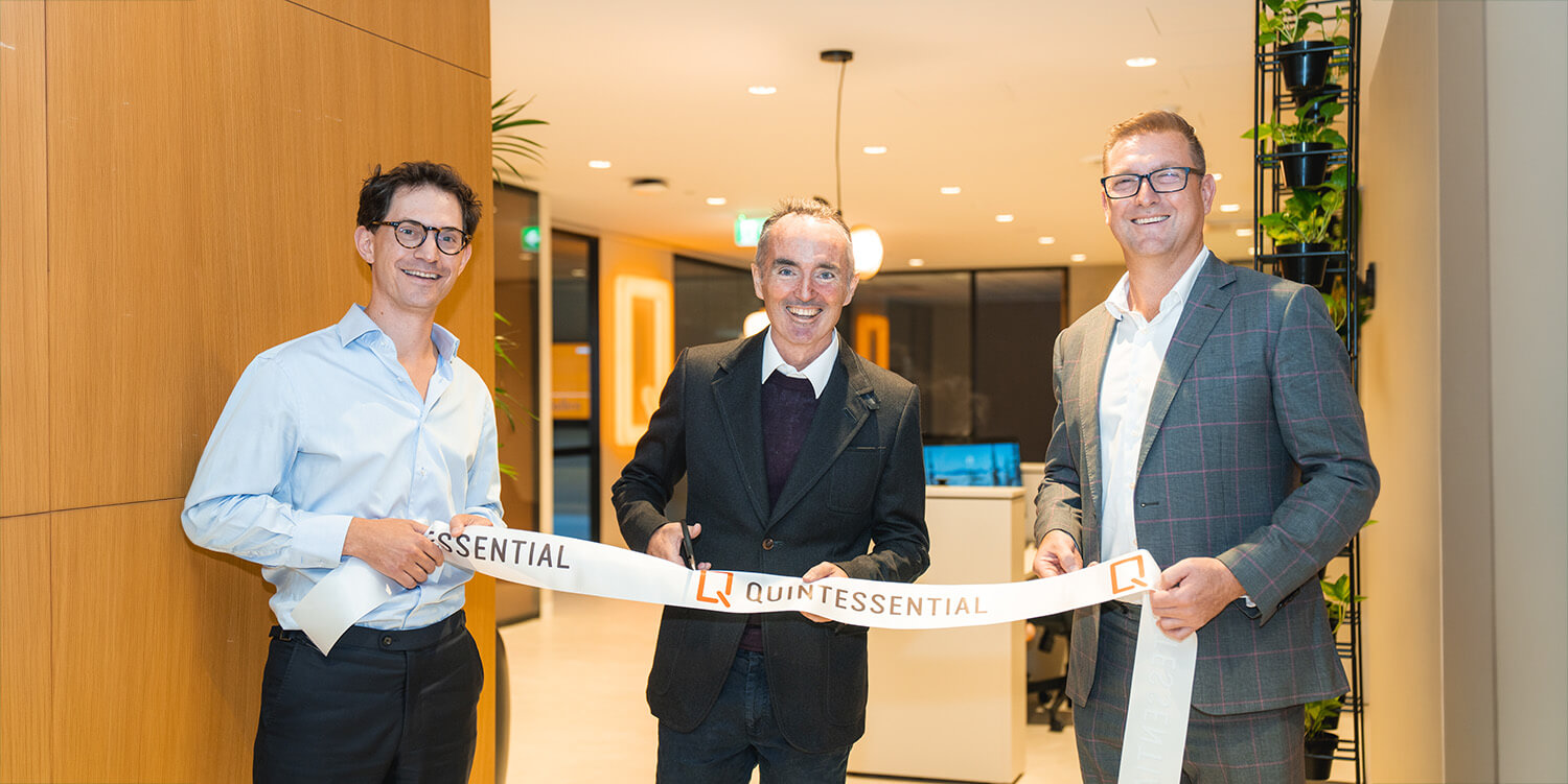 Three men smiling and holding a white ribbon that reads Quintessential during a ribbon-cutting ceremony.