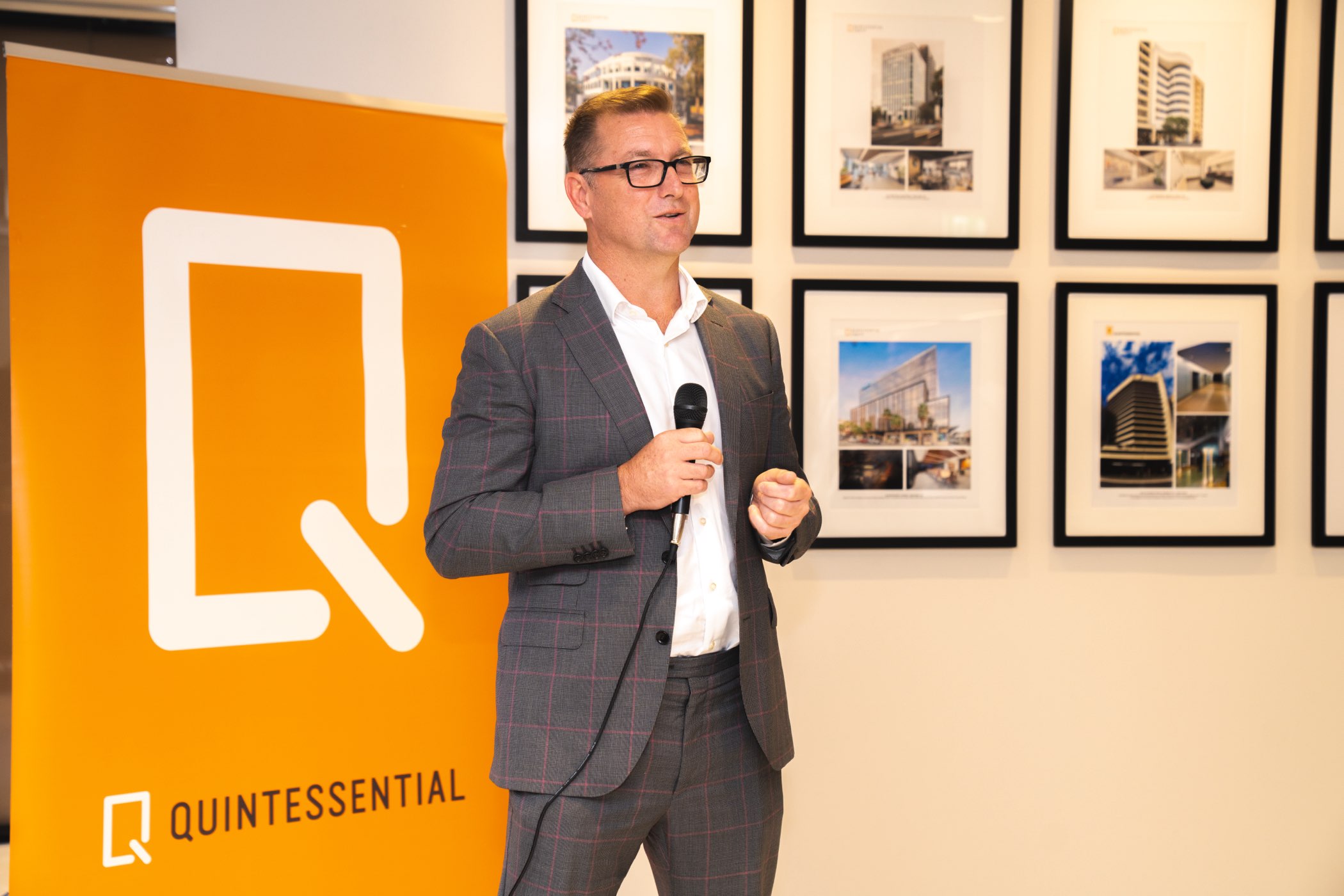 Man in a gray suit and glasses speaking into a microphone in front of an orange Quintessential banner and framed building photos on a wall.