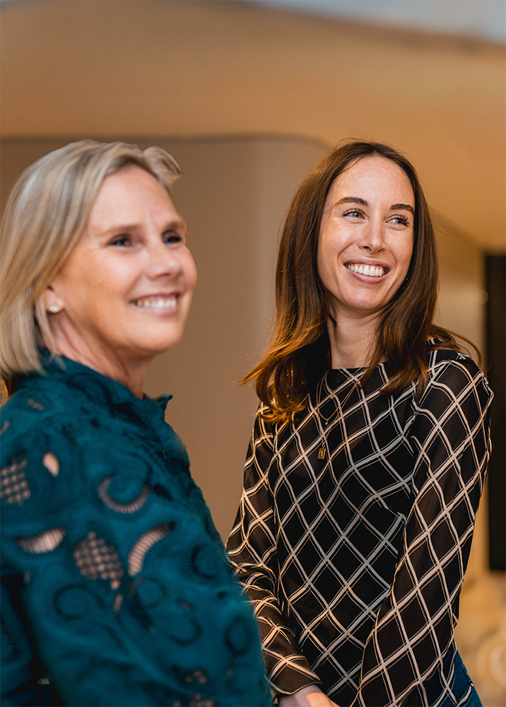 Two women smiling and looking to the side in a warmly lit indoor setting.