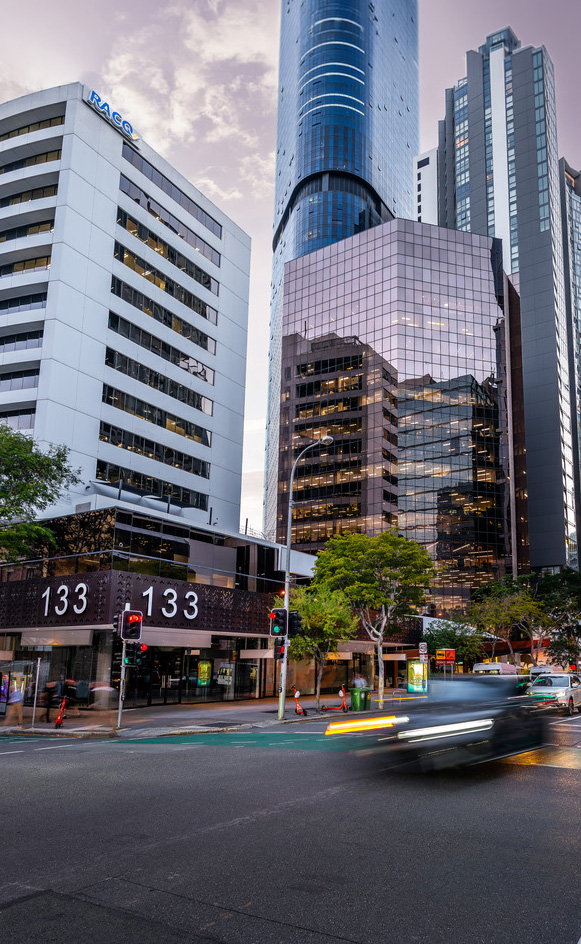 City street with modern tall buildings, traffic lights, trees, and blurred car headlights at dusk.