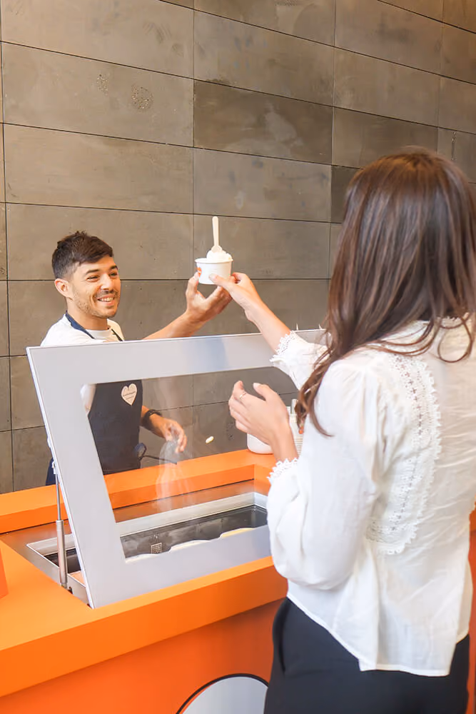Man behind counter hands cup of ice cream to woman in white blouse at ice cream shop.