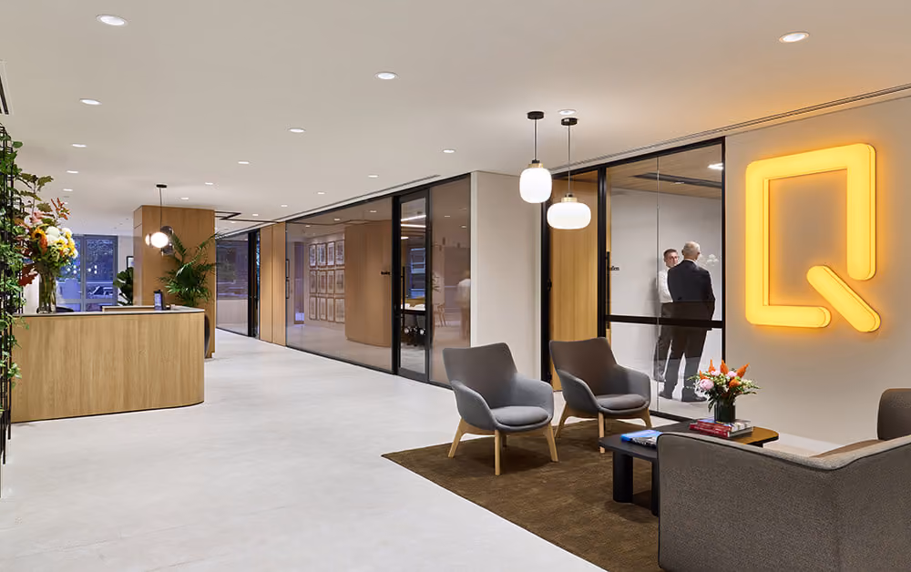 Modern office lobby with wooden reception, gray chairs, glass meeting rooms, and two men talking inside.
