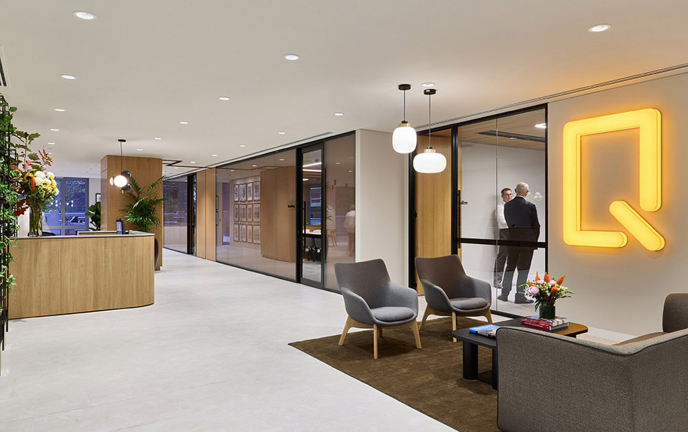 Modern office lobby with wooden reception, gray chairs, glass meeting rooms, and two men talking inside.