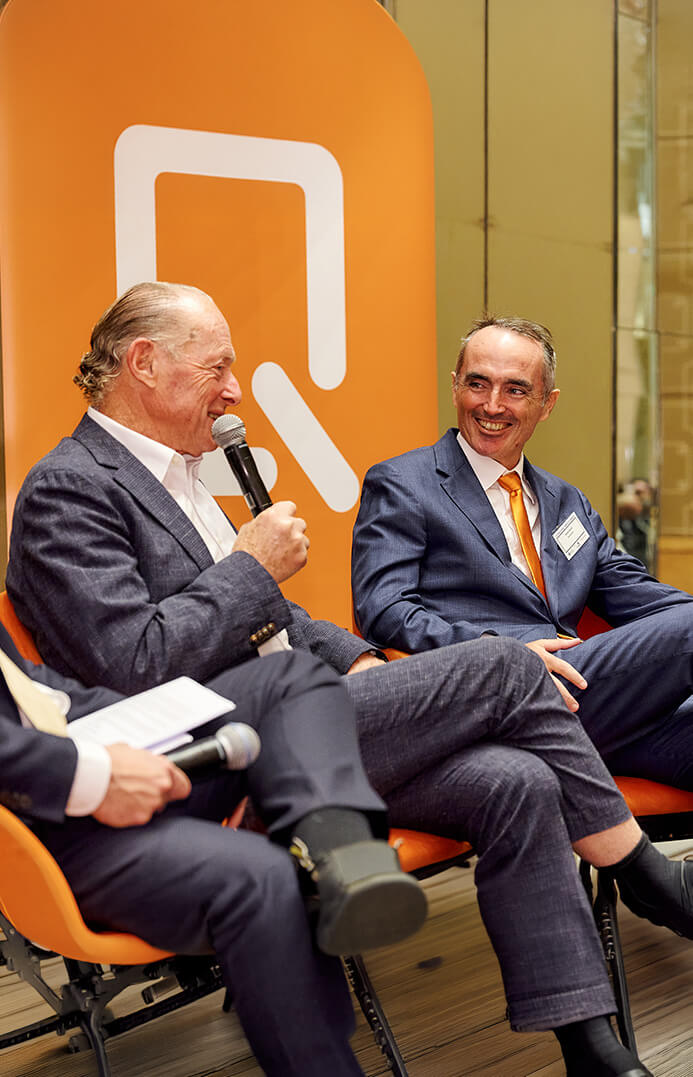 Two men in suits sitting and talking with microphones in front of an orange backdrop with a white logo.