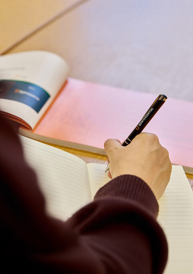 Person writing in a lined notebook with a black QUINTESSENTIAL pen on a desk with books.
