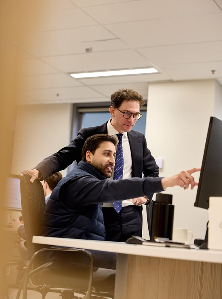 Two men in an office looking at a computer screen, one sitting and pointing, the other standing.