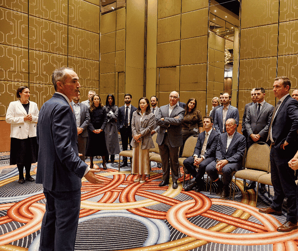 Man in suit speaking to seated and standing business people in a modern conference room.