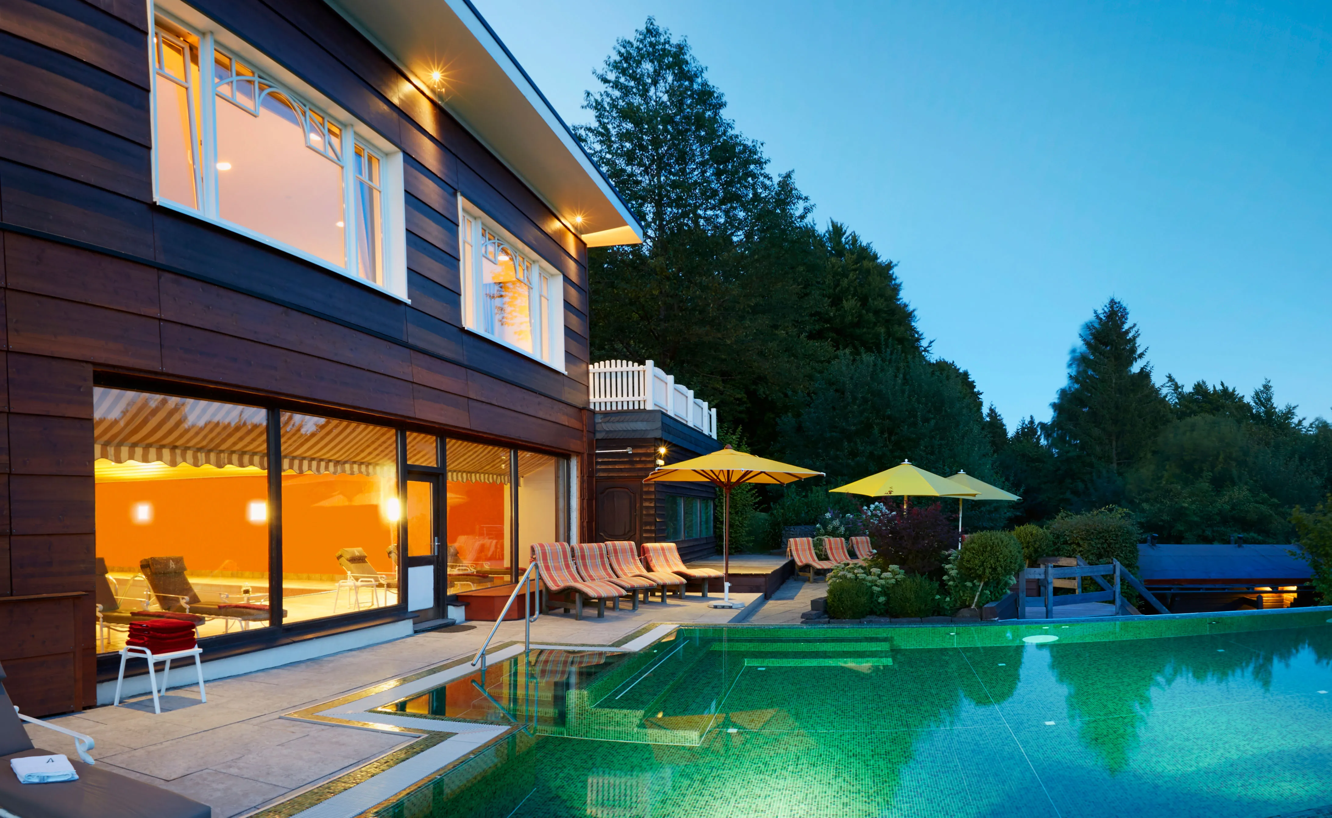 Außenpool mit Liegestühlen und gelben Sonnenschirmen neben einem beleuchteten Holzhaus bei Dämmerung, umgeben von Bäumen.