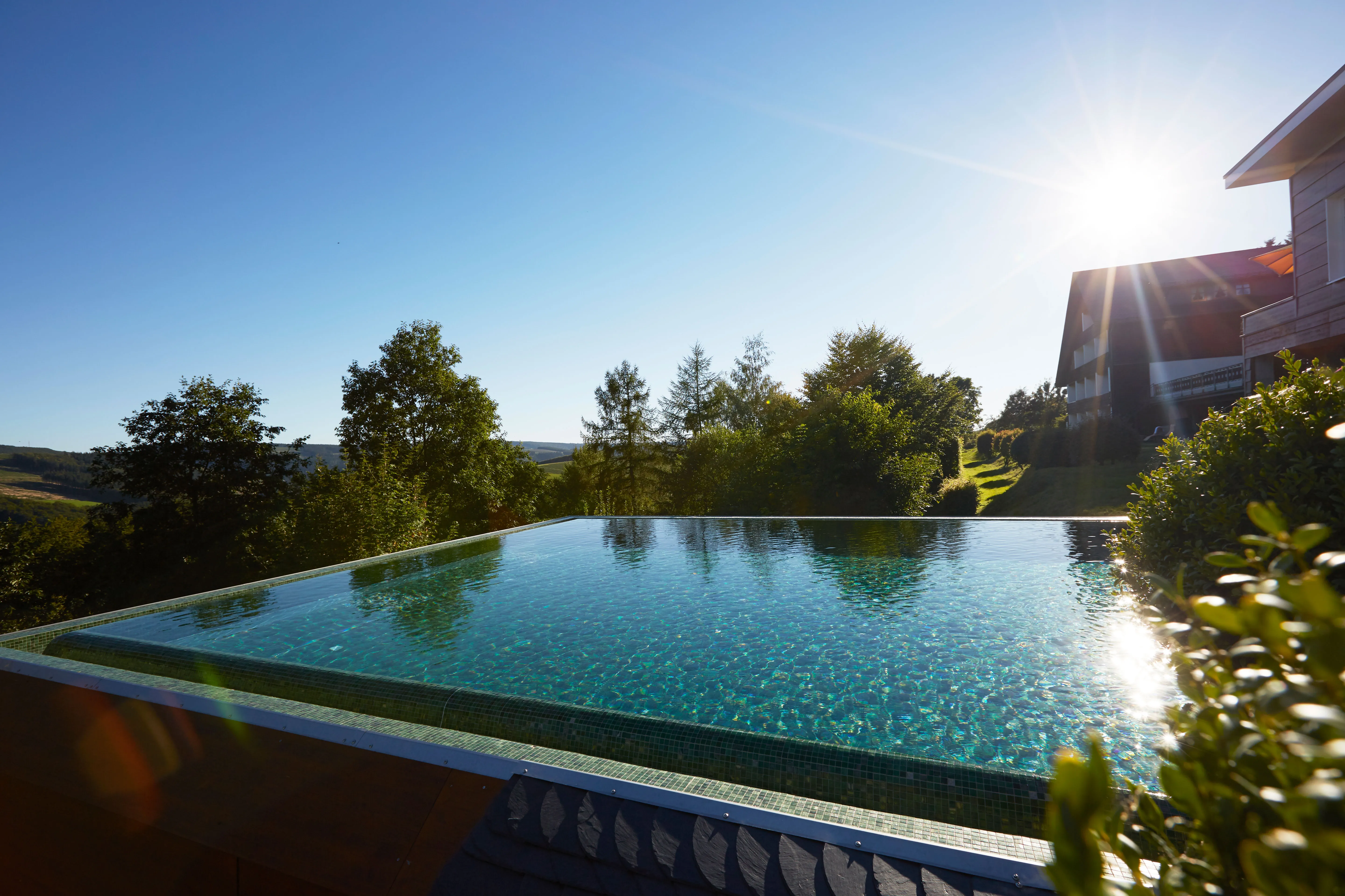 Infinity-Pool mit Blick auf bewaldete Hügel und Sonne am klaren Himmel.