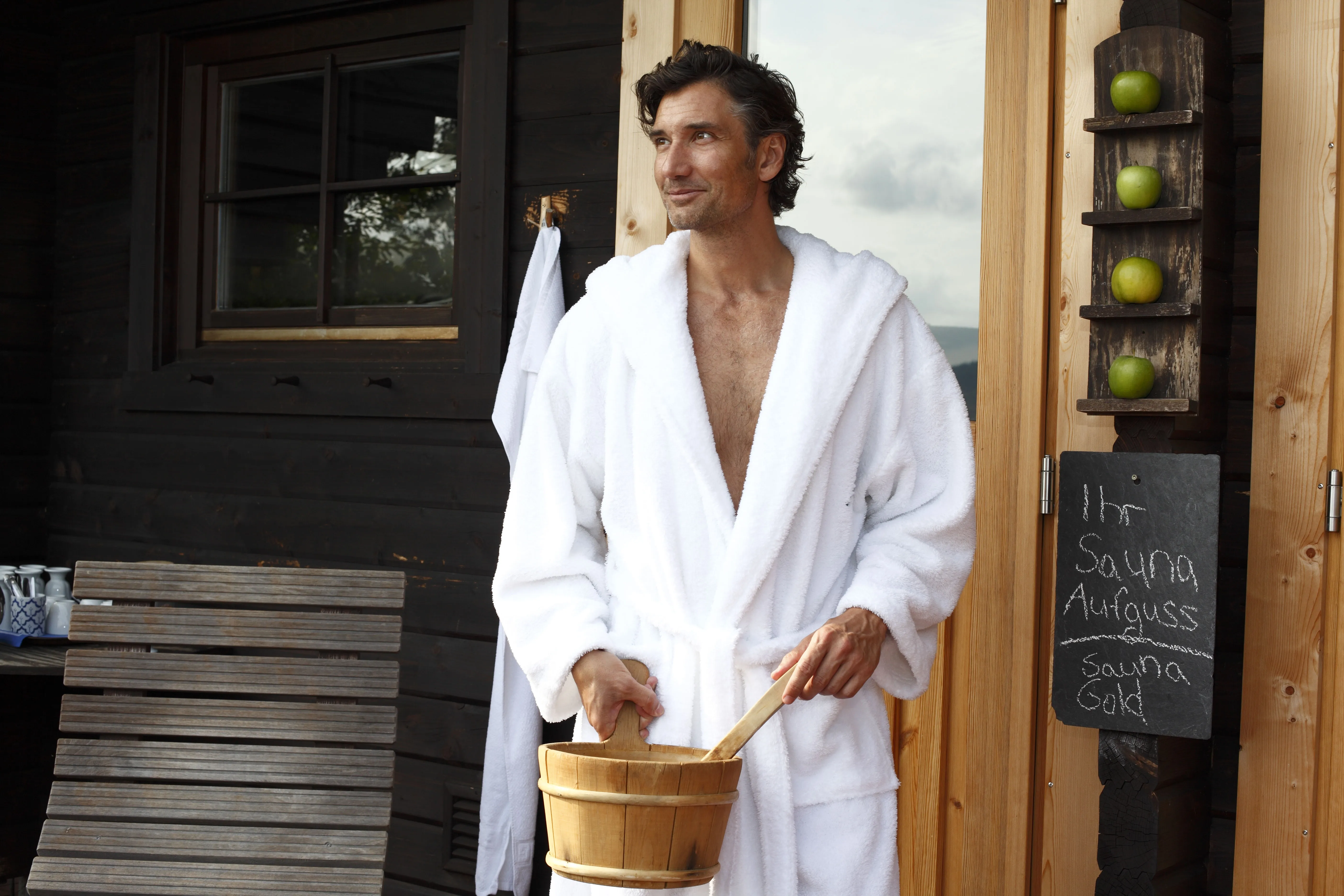 Mann in weißem Bademantel hält einen Holzeimer vor einer Saunatür mit einem Schild und vier grünen Äpfeln.