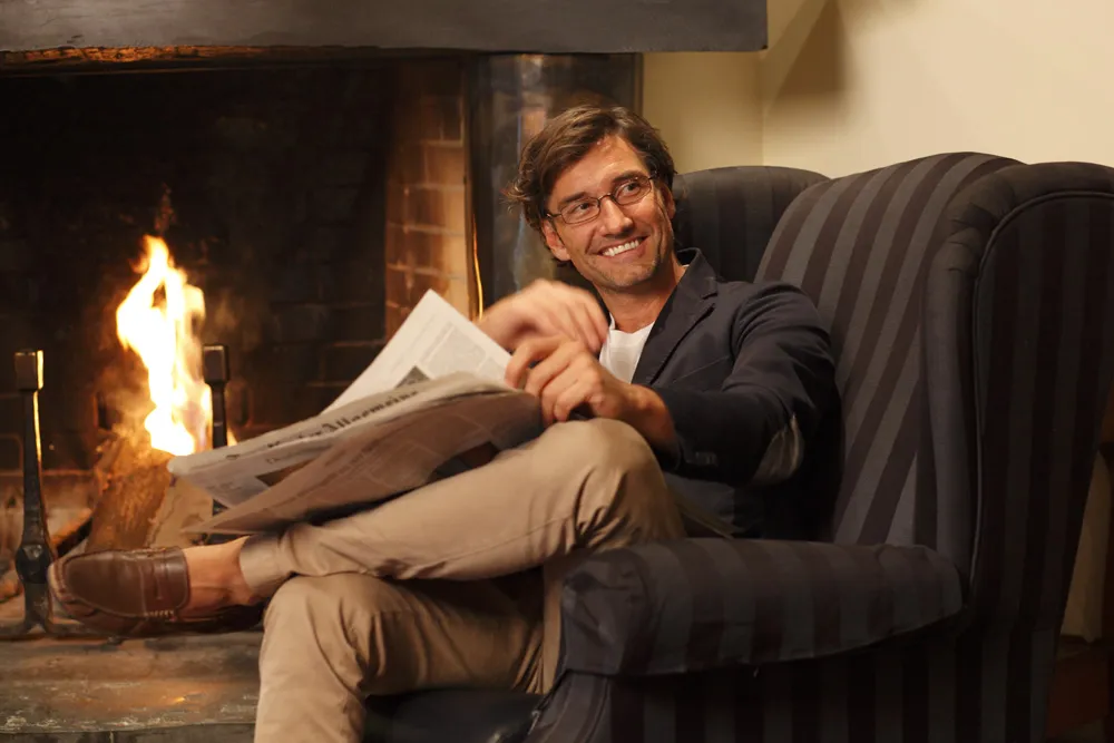 Mann sitzt entspannt in einem gestreiften Sessel vor einem Kamin und liest eine Zeitung.