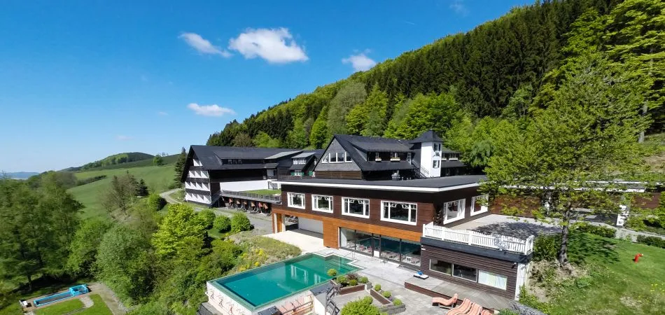Modernes Hotel mit Schwimmbad im Außenbereich, umgeben von grünen Bäumen und Hügeln unter blauem Himmel.
