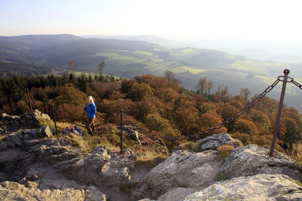 Frau mit blauer Jacke wandert auf felsigem Pfad mit Kettengeländer, Blick auf herbstliche Wälder und sanfte Hügel im Hintergrund.
