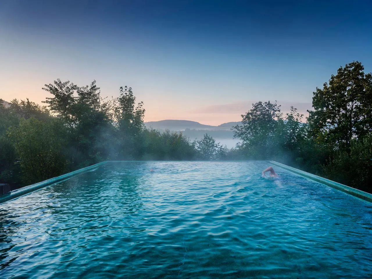 Ein Schwimmer im beheizten Infinity-Pool mit Blick auf bewaldete Hügel und einen klaren Morgenhimmel.
