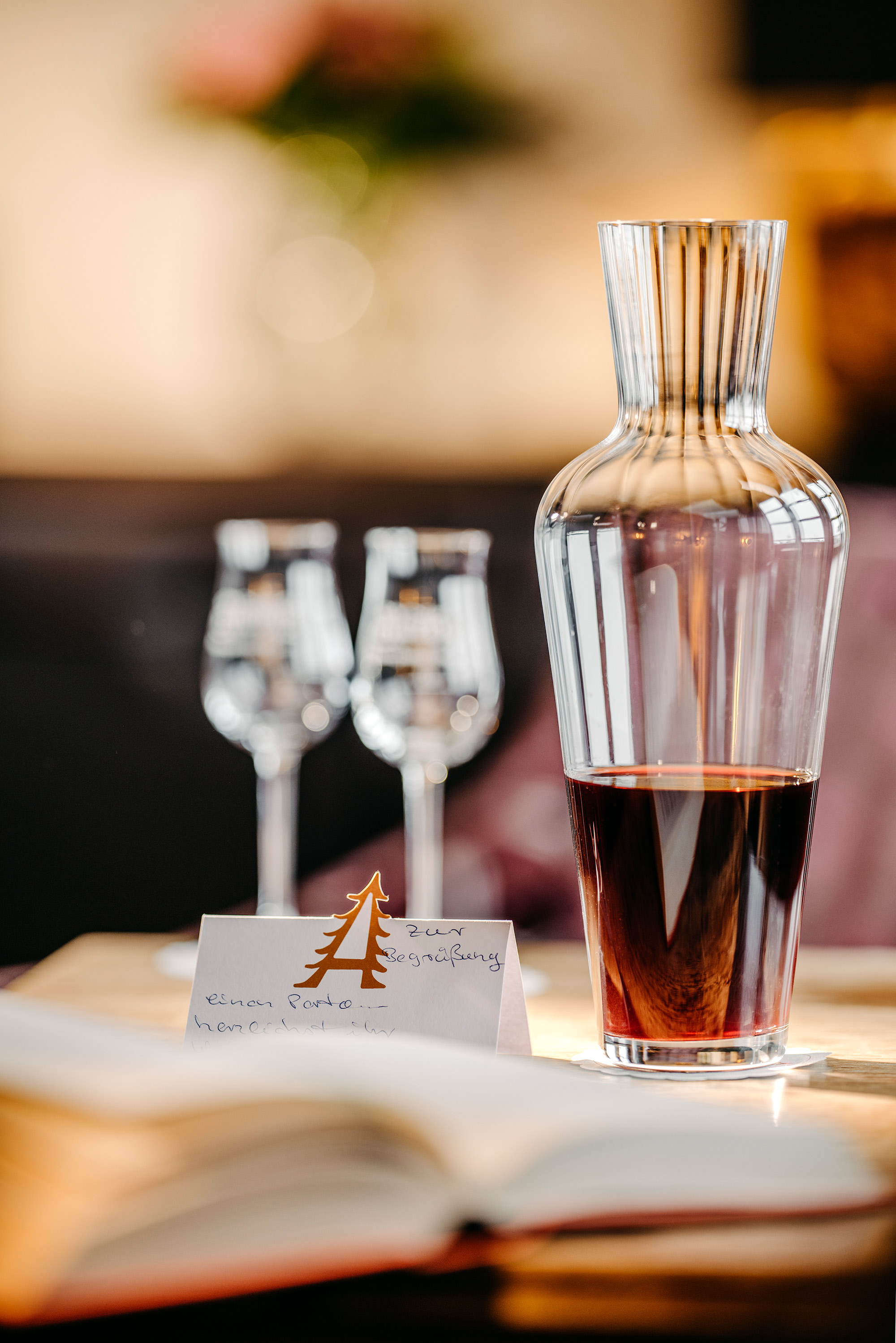 Glaskaraffe mit rotem Getränk, zwei Weingläser im Hintergrund und eine beschriebene Platzkarte auf einem Holztisch.