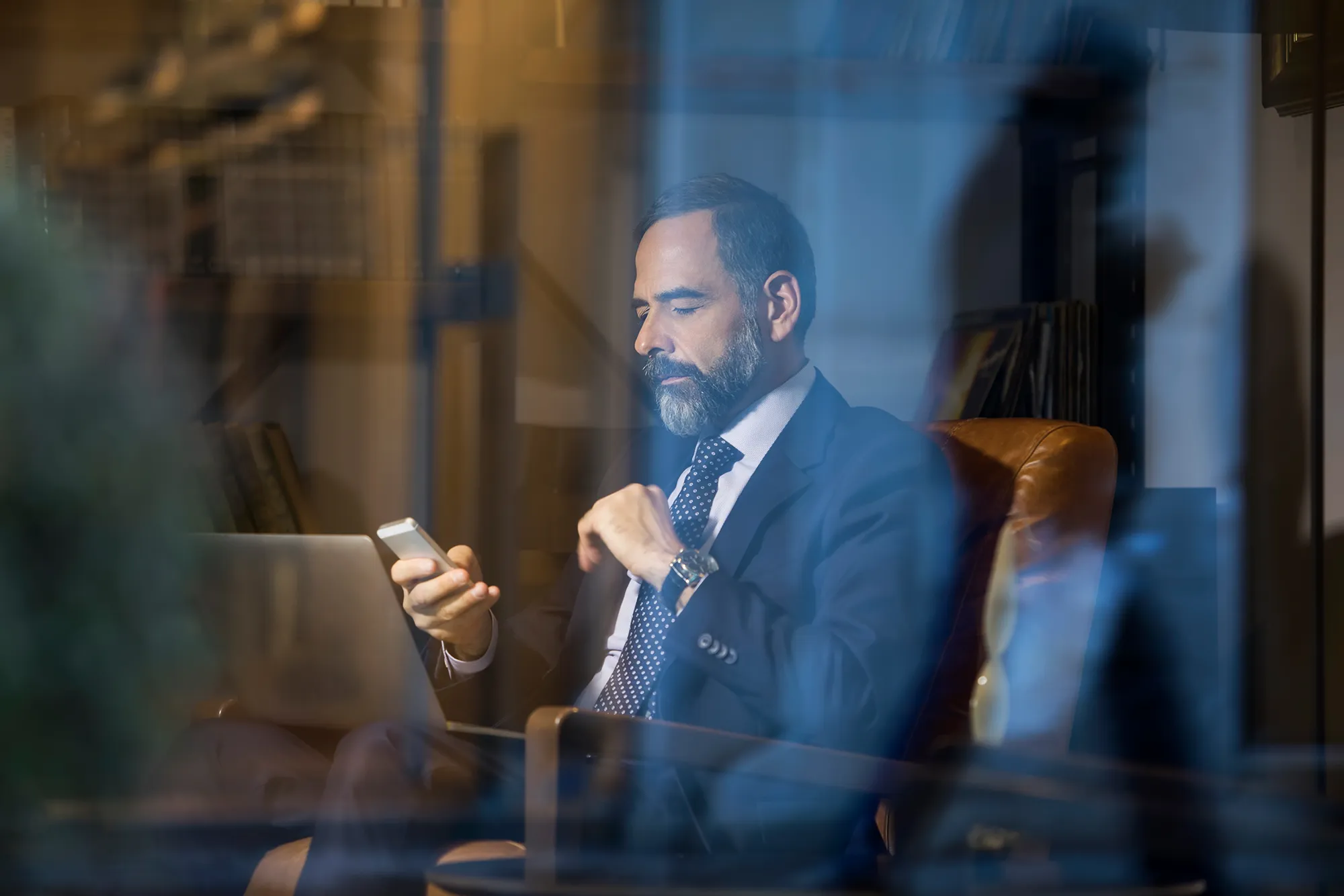 Geschäftsmann im Anzug sitzt in einem Ledersessel und blickt auf sein Smartphone, mit einem Laptop auf dem Schoß.