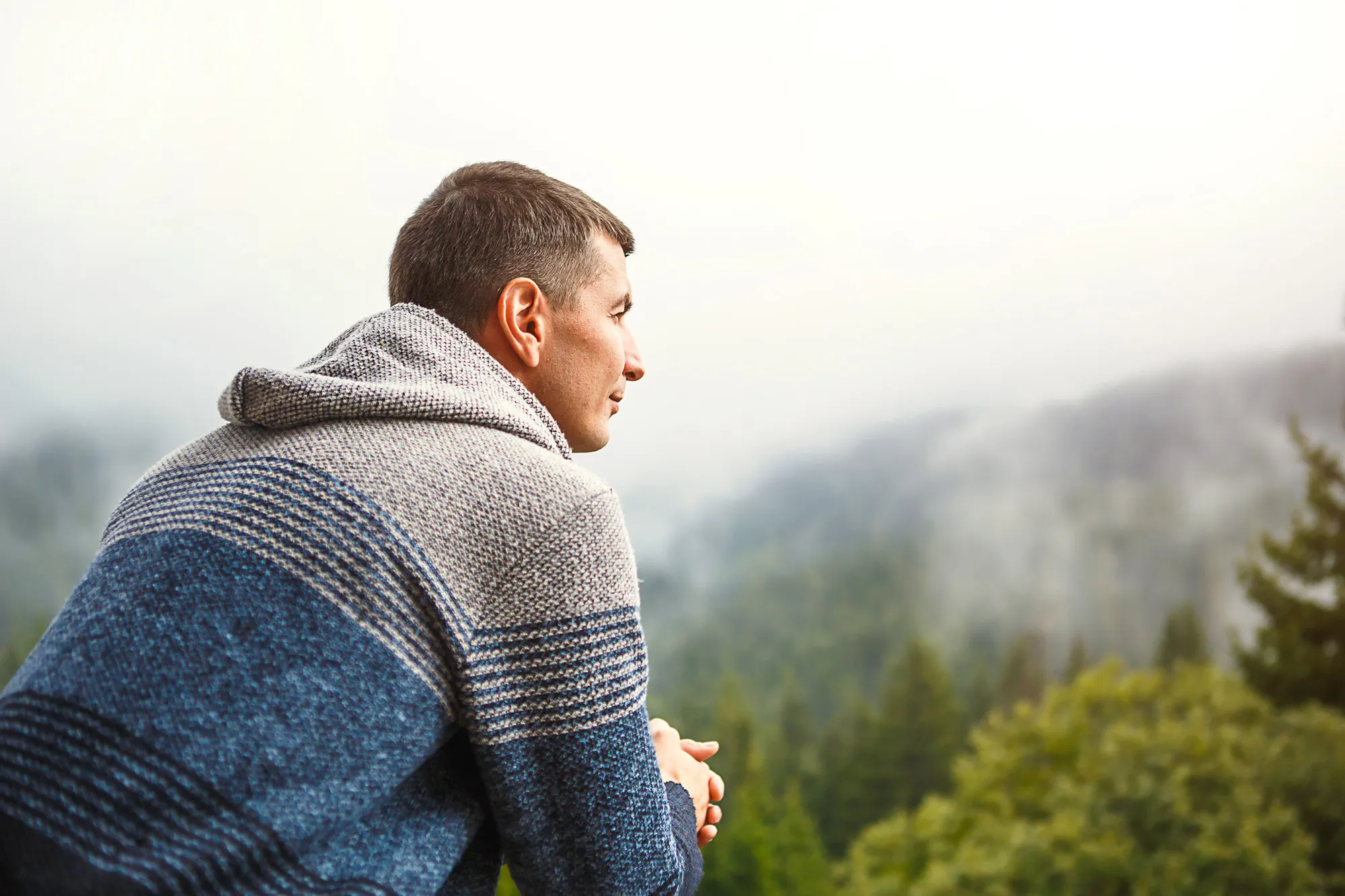 Mann in grauem und blauem Pullover blickt nachdenklich in neblige bewaldete Berge.