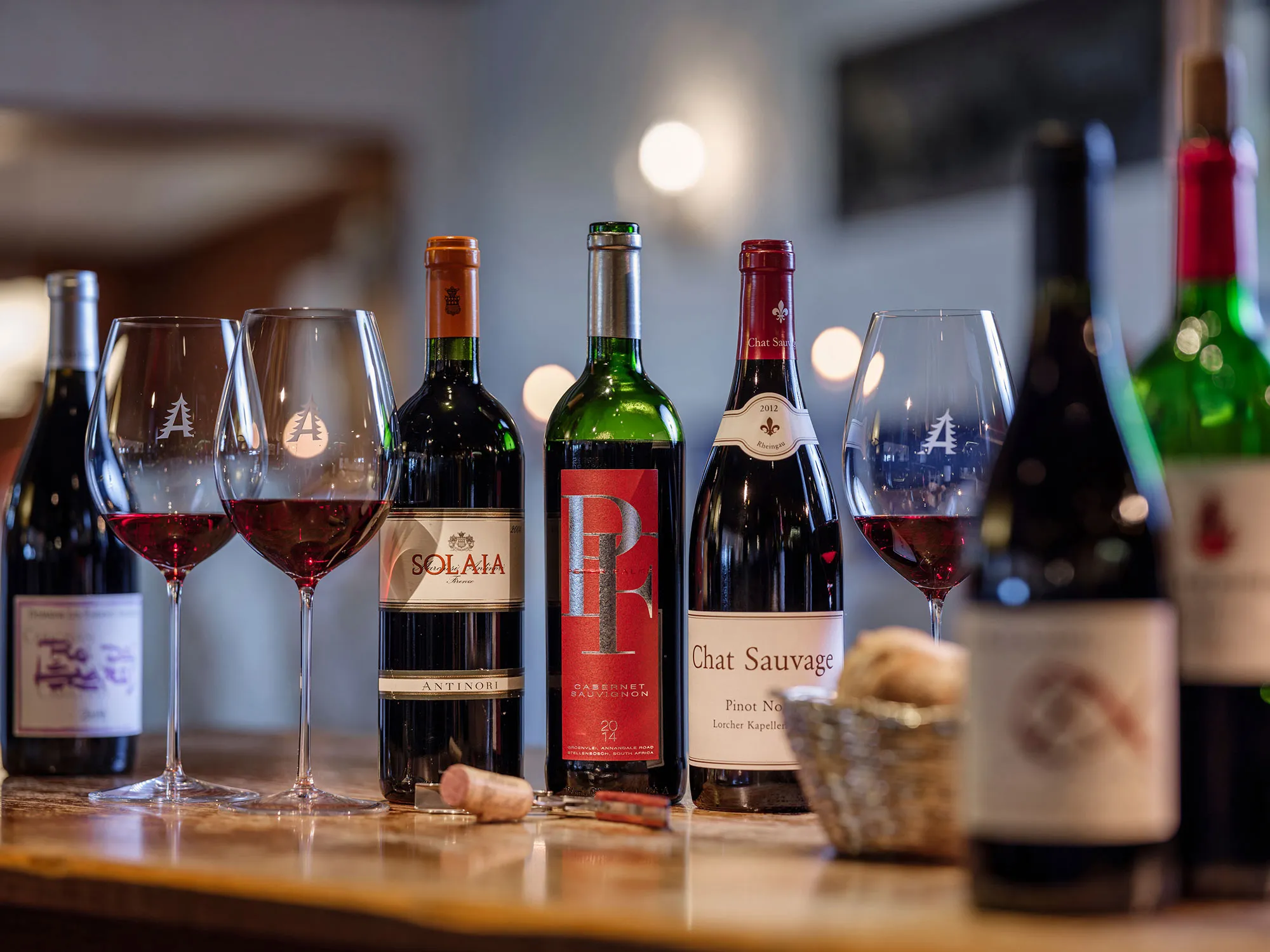 Mehrere Rotweinflaschen und drei teilweise gefüllte Weingläser auf einem Holztisch in einem gemütlichen Raum.