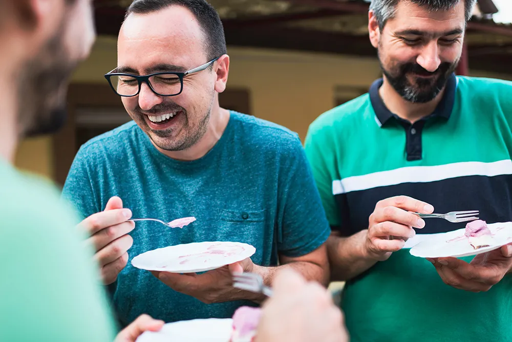 Drei Männer lachen und essen zusammen Kuchen auf Papptellern im Freien.