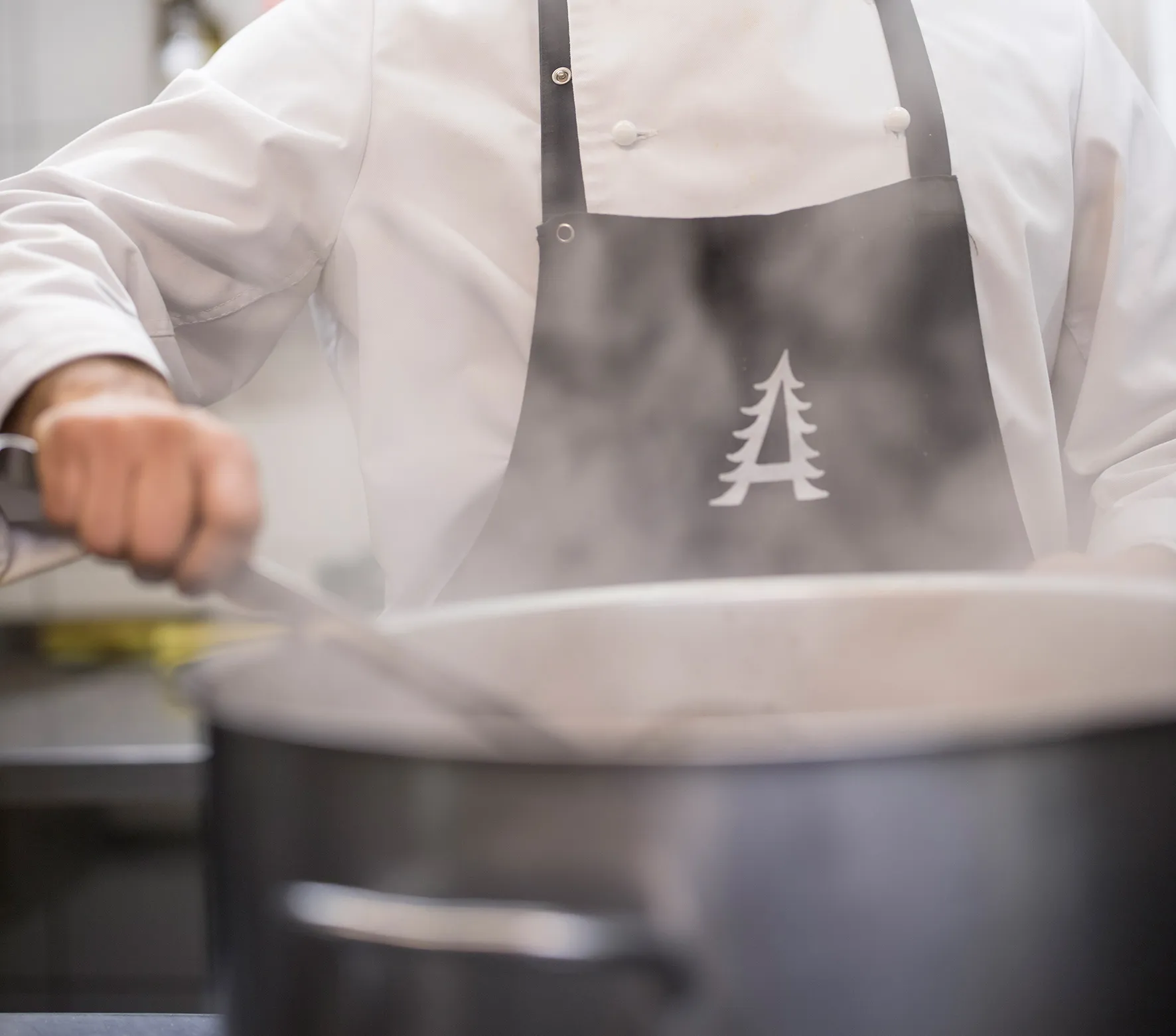 Koch in weißer Uniform und schwarzer Schürze rührt einen großen dampfenden Topf.
