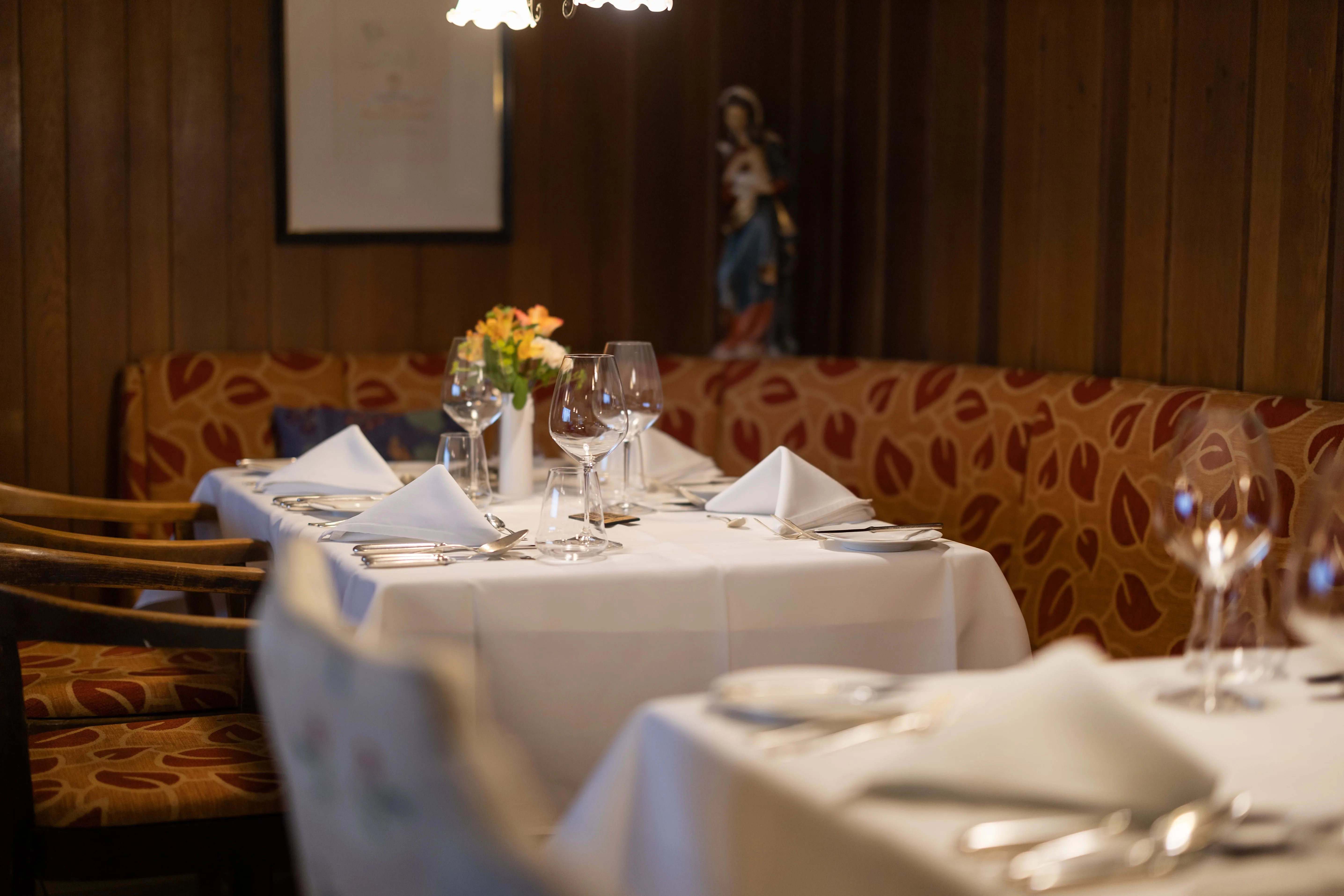 Eleganter Esstisch in einem Restaurant mit weißen Tischdecken, gefalteten Servietten, Weingläsern und einer Blumenvase, umgeben von gemusterten Polstersesseln.
