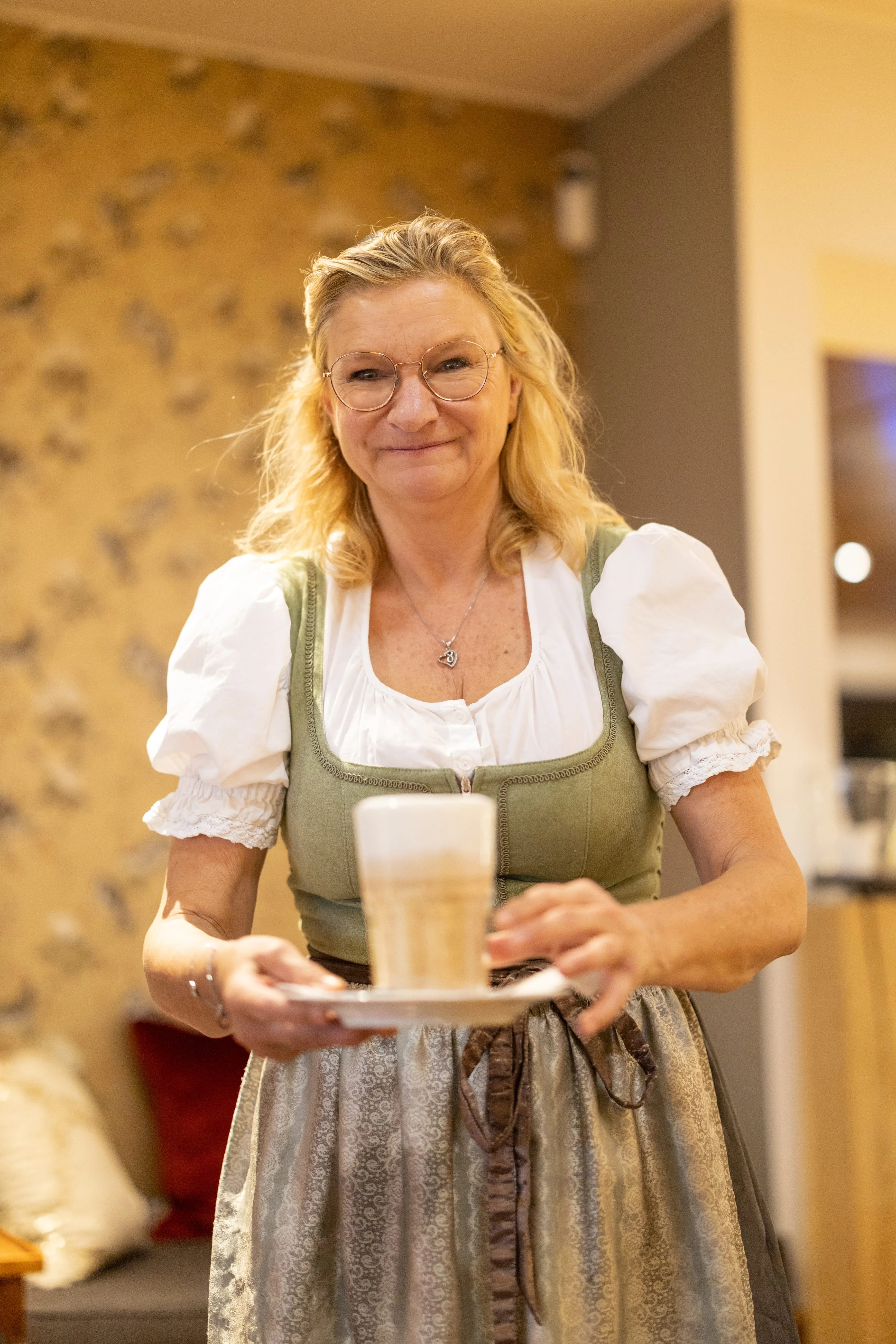 Frau im traditionellen Dirndl mit Brille lächelt und hält ein Latte Macchiato auf einer Untertasse.