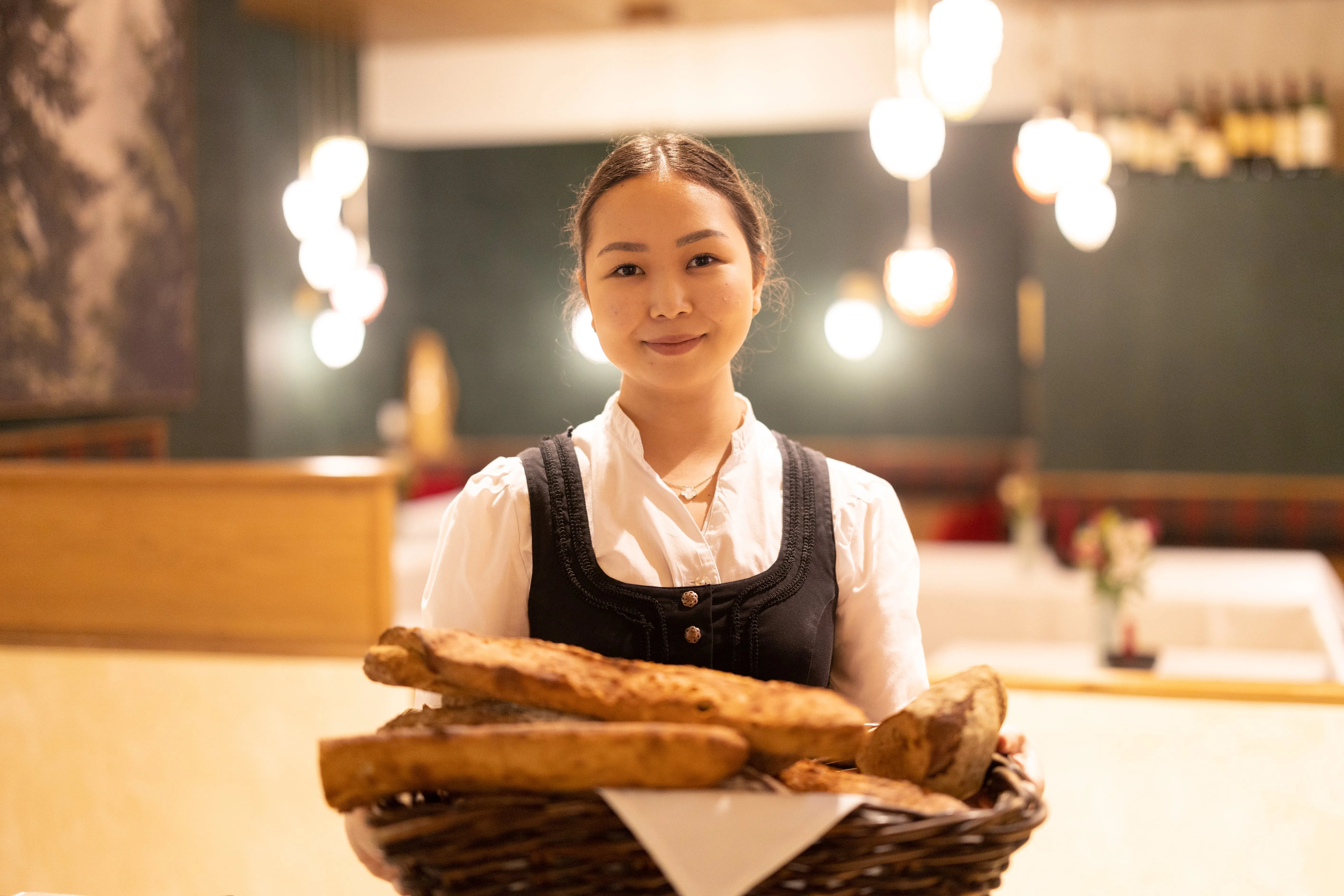 Junge Kellnerin im traditionellen Dirndl hält einen Korb mit frisch gebackenem Brot in einem Restaurant.