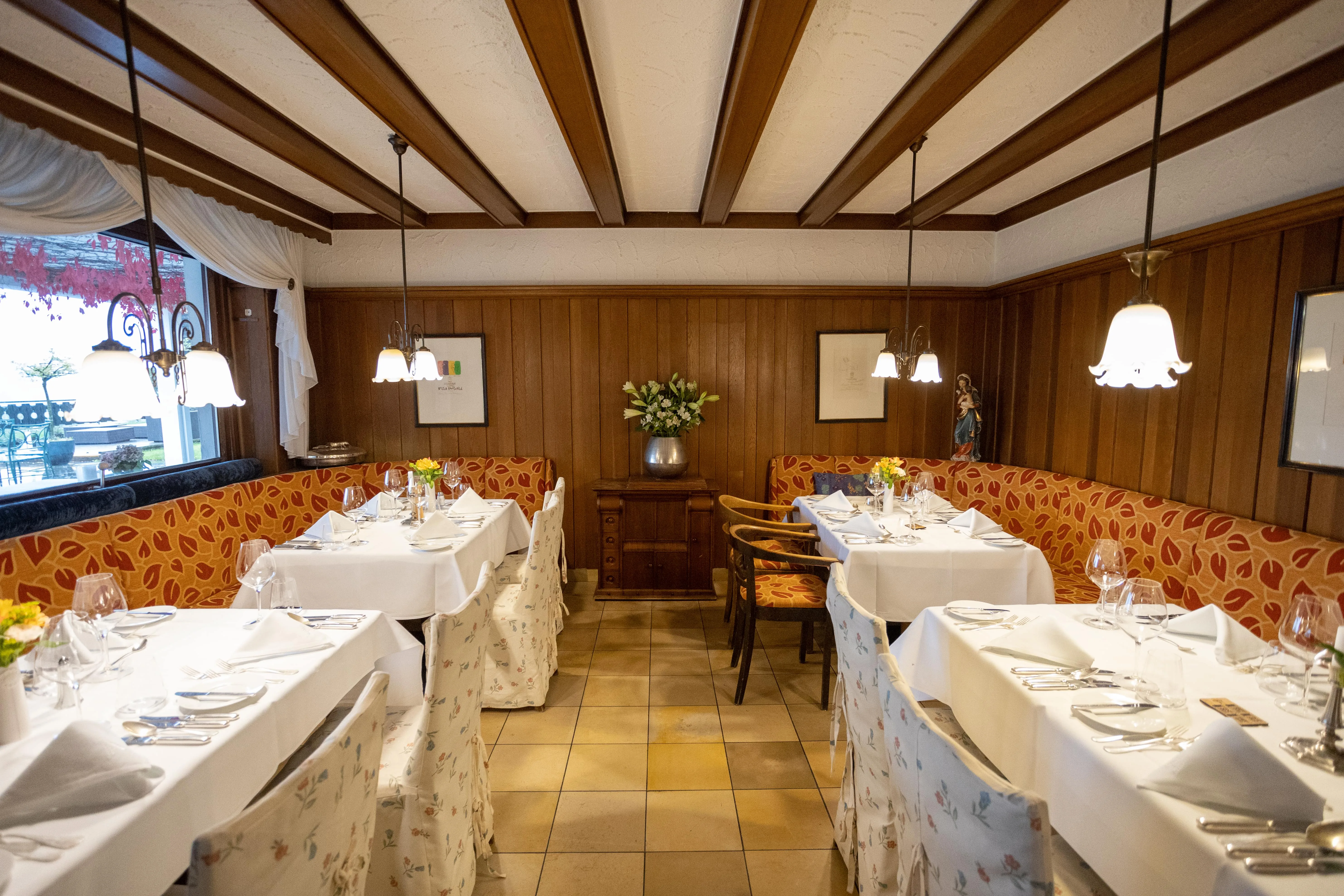 Gemütliches Restaurant mit Holzwänden, weißen Tischdecken, floral bezogenen Stühlen und herabhängenden Lampen.