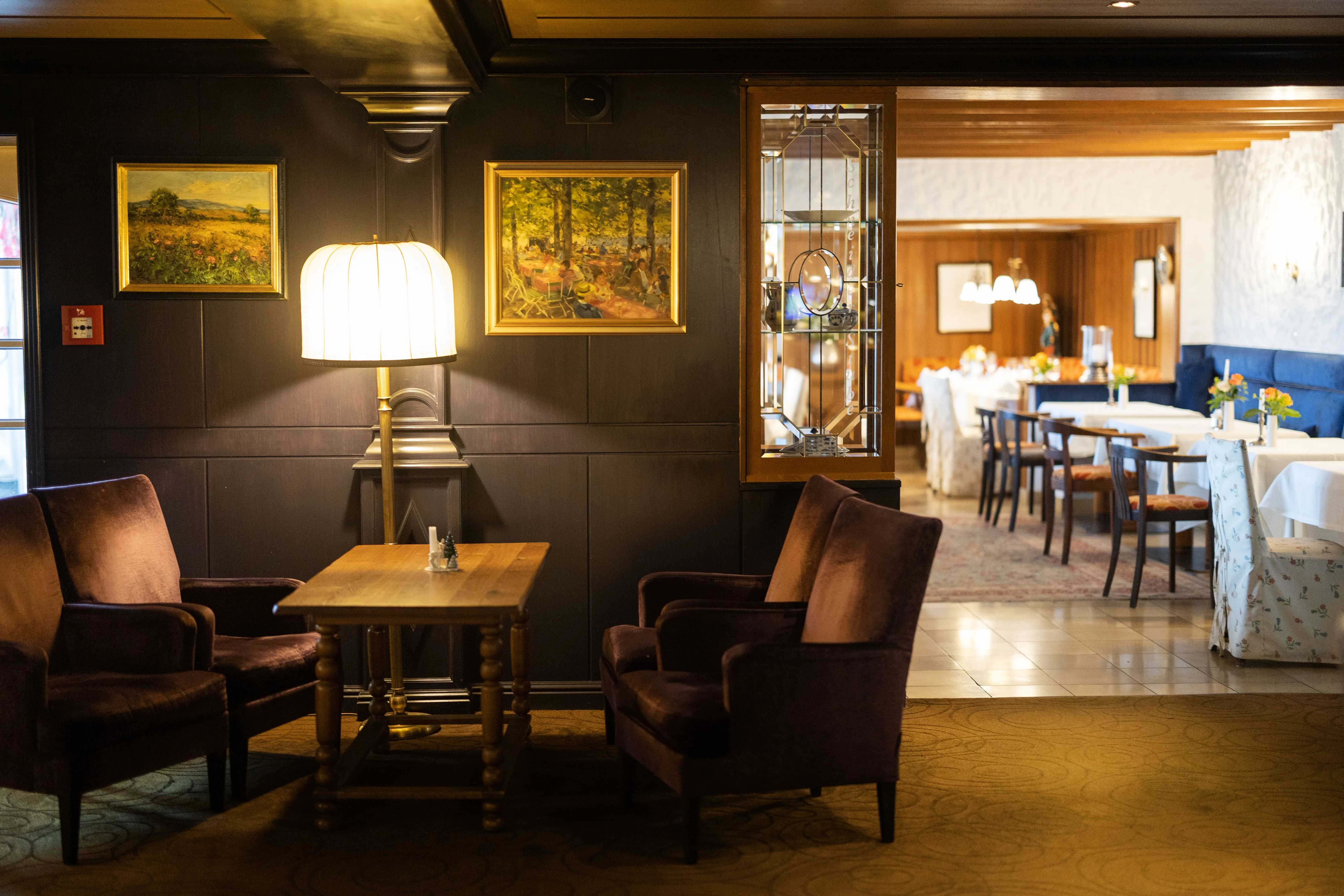 Gemütlicher Sitzbereich mit zwei braunen Sesseln, einem Holztisch und einer Stehlampe vor dunkler Wand mit zwei Landschaftsbildern, daneben Essbereich mit gedeckten Tischen und Stühlen.