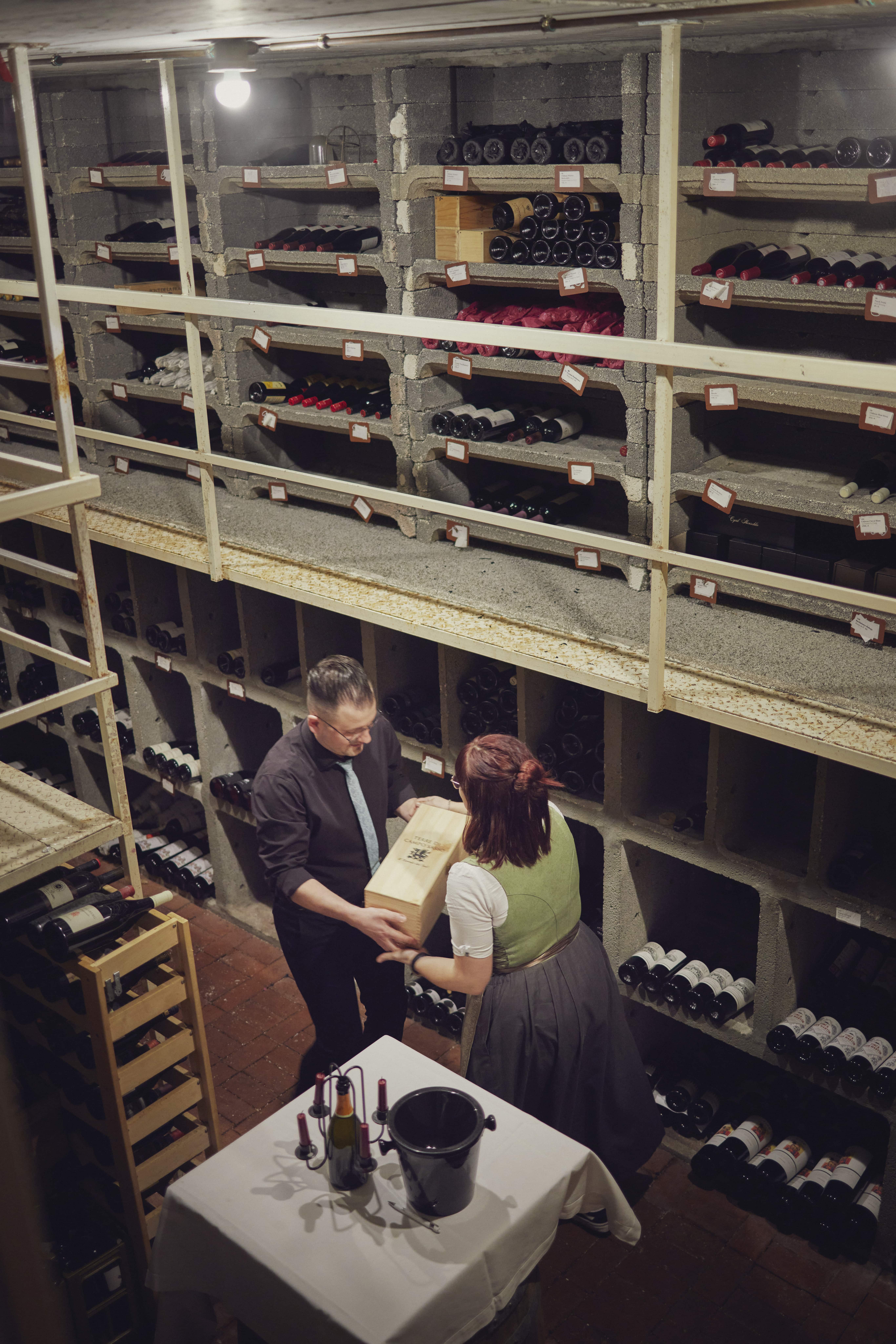 Zwei Personen übergeben sich in einem Weinkeller eine Holzkiste mit Weinflaschen, umgeben von Weinregalen.