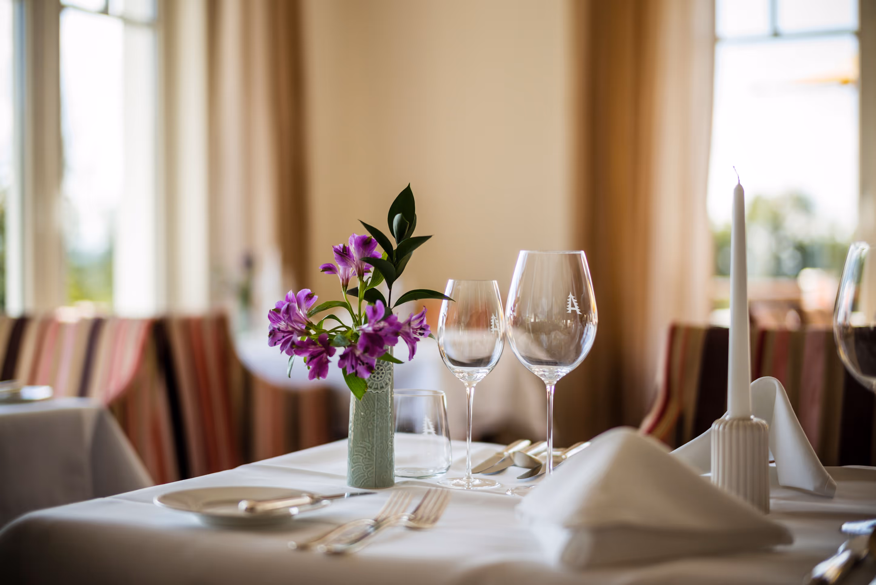 Eleganter gedeckter Tisch mit weißen Servietten, zwei Weingläsern, einer Vase mit lila Blumen und einer weißen Kerze in einem Restaurant.
