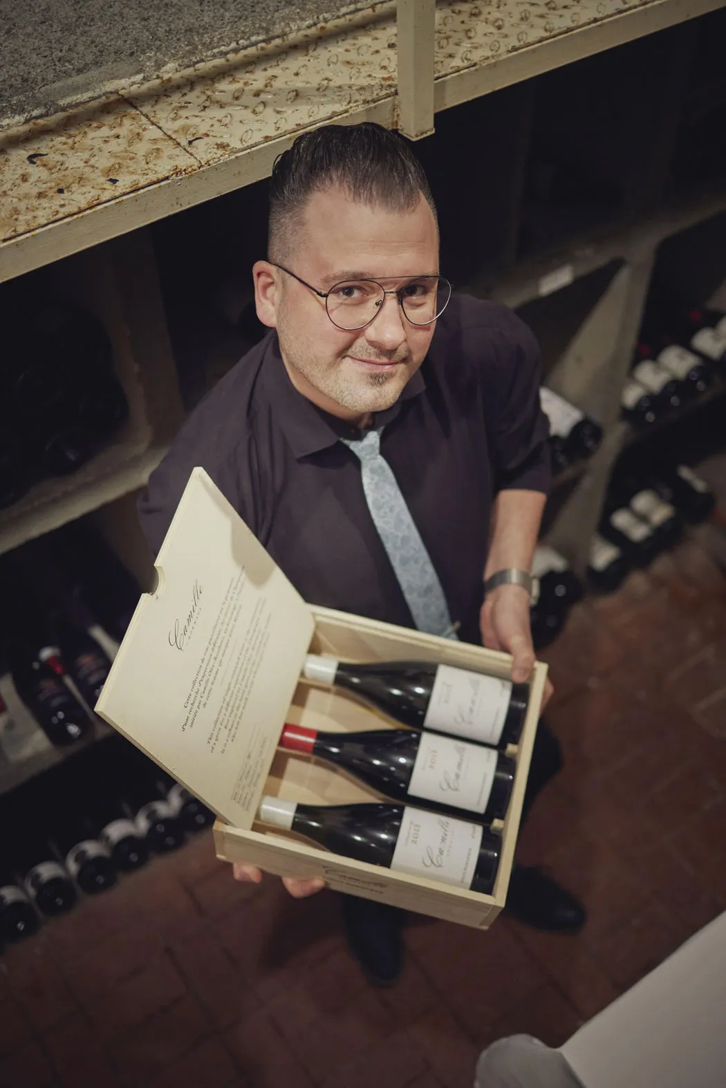 Mann mit Brille in einem Weinkeller zeigt eine geöffnete Holzkiste mit drei Weinflaschen.