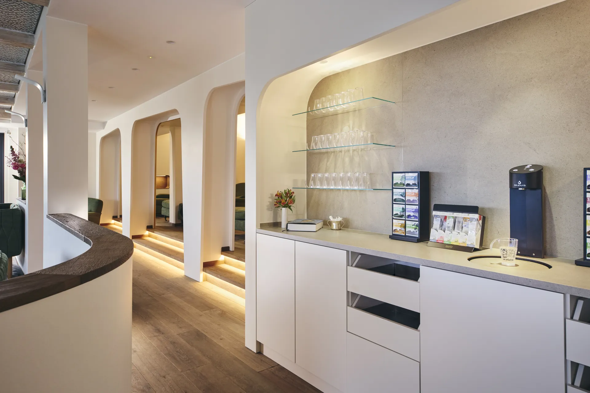 Moderne, minimalistische Küchenzeile mit weißen Schränken, zwei offenen Schubladen, Glasregalen und schwarzem Wasserhahn unter eingebauten Deckenleuchten.