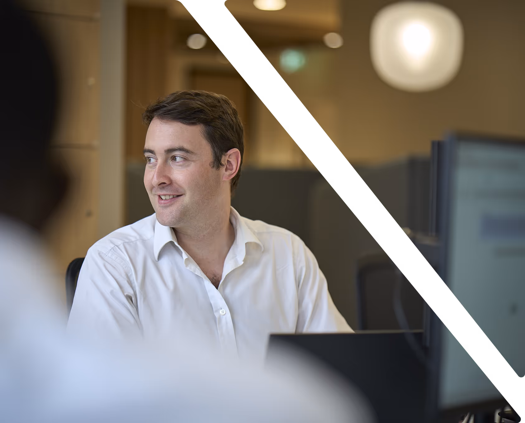 Smiling man in a white shirt looking to the right while seated at a desk with a computer monitor, with diagonal black bars partially overlaying.