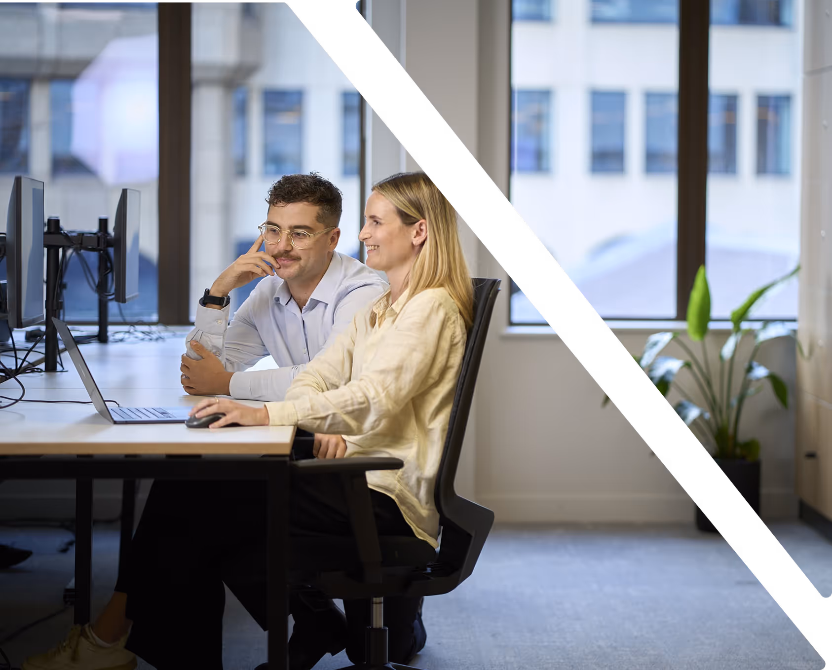 Two colleagues, a man and a woman, sitting side-by-side and smiling while viewing a laptop screen in an office, with diagonal black bars partially overlaying the image.