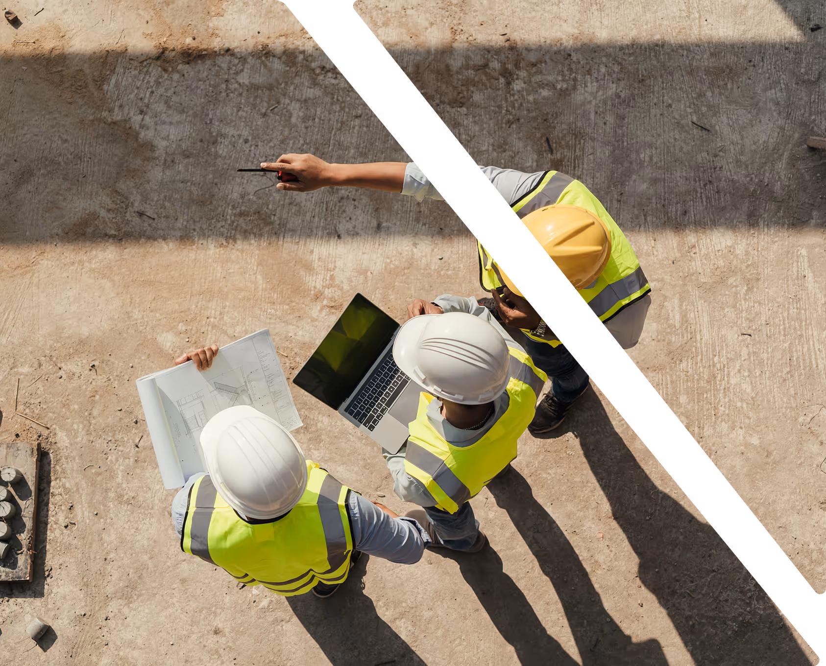 Aerial view of three construction workers wearing hard hats and reflective vests reviewing a blueprint and laptop at a worksite.