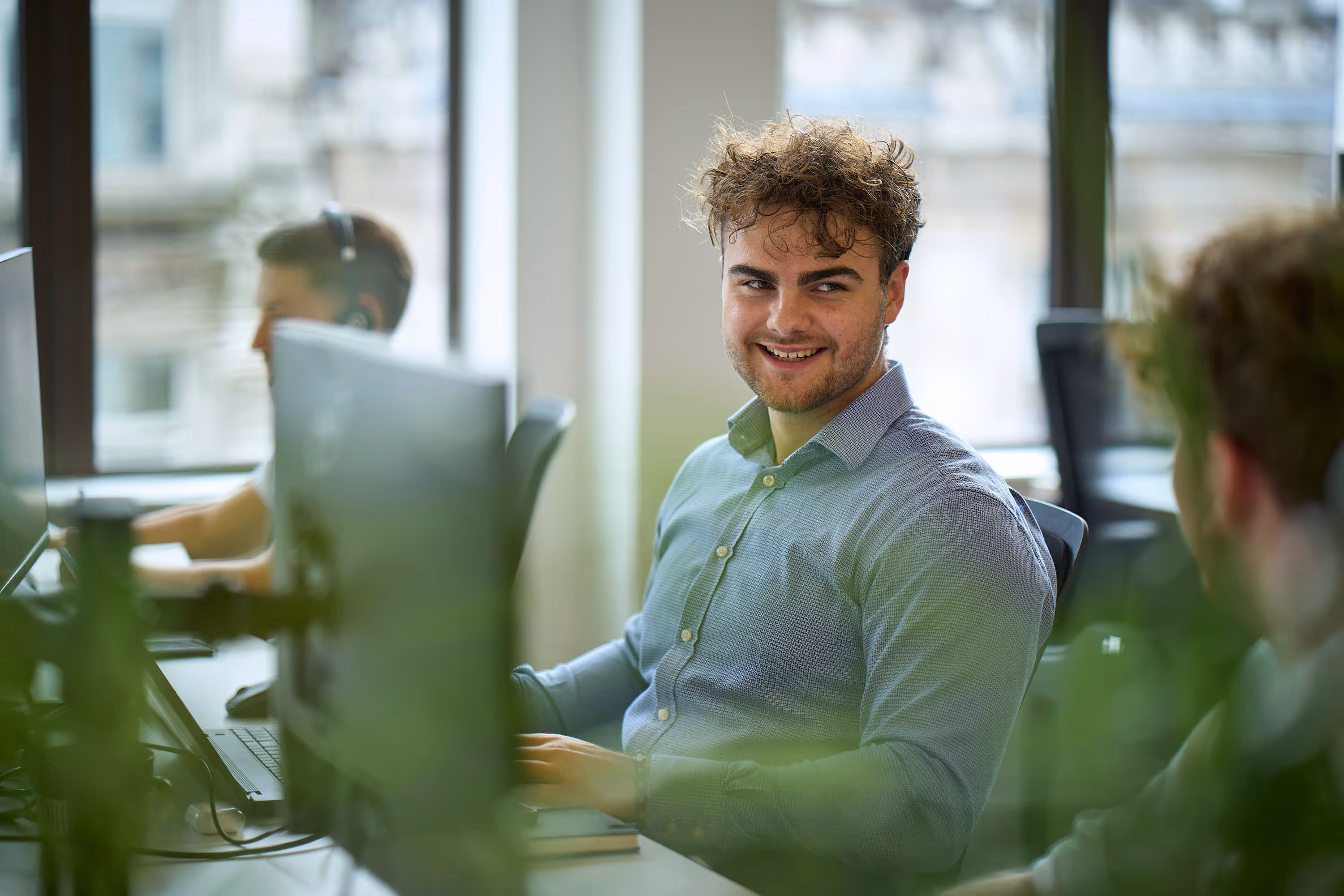 Young man with curly hair in a light blue shirt smiling and talking to a colleague in an office.