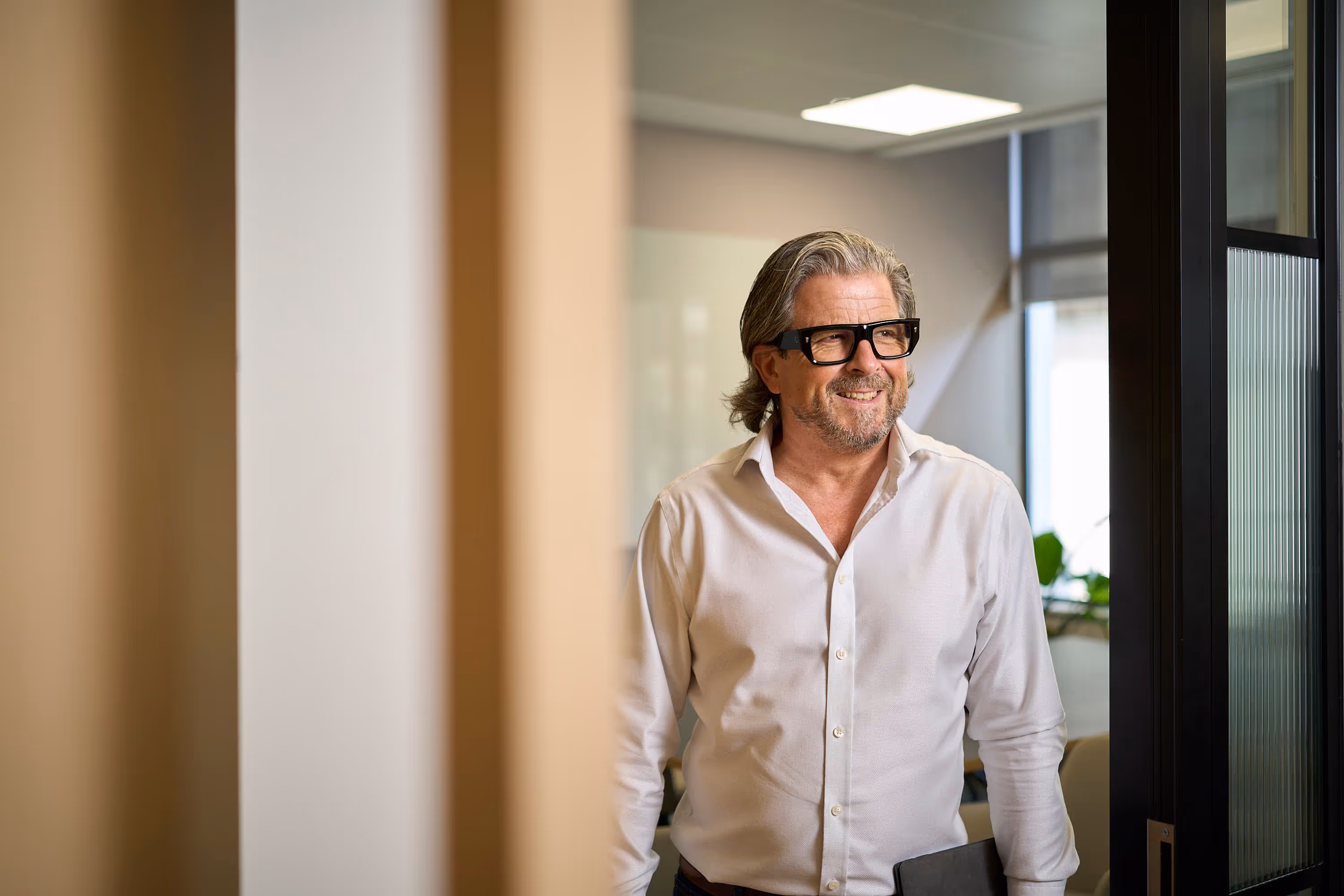 Man with glasses and beard smiling in a white shirt in a brightly lit office near a window.