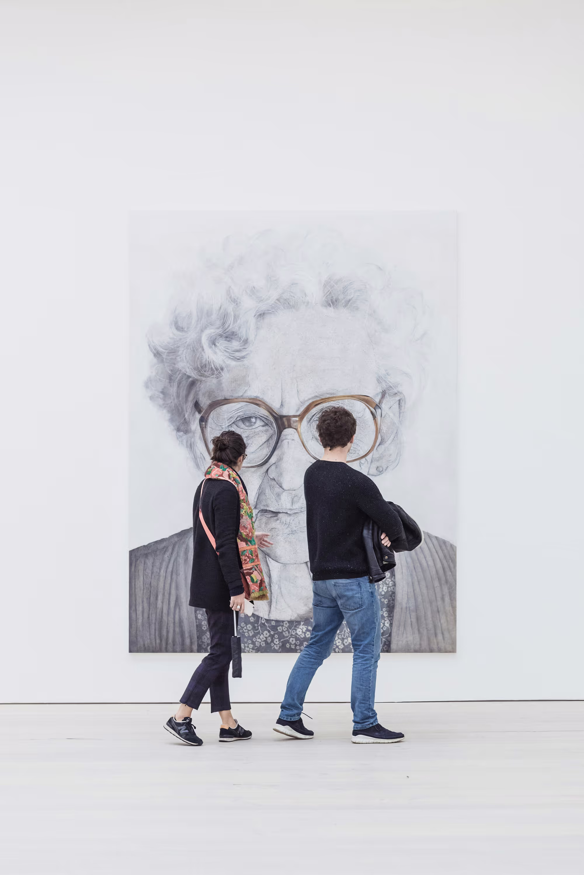 Two people observing a large portrait of an elderly woman in an art gallery