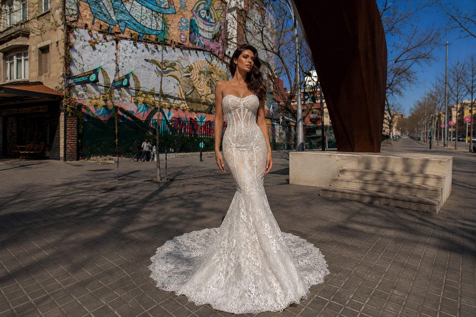 Donna in abito da sposa bianco aderente con strascico, posa su strada urbana con graffiti di sfondo.