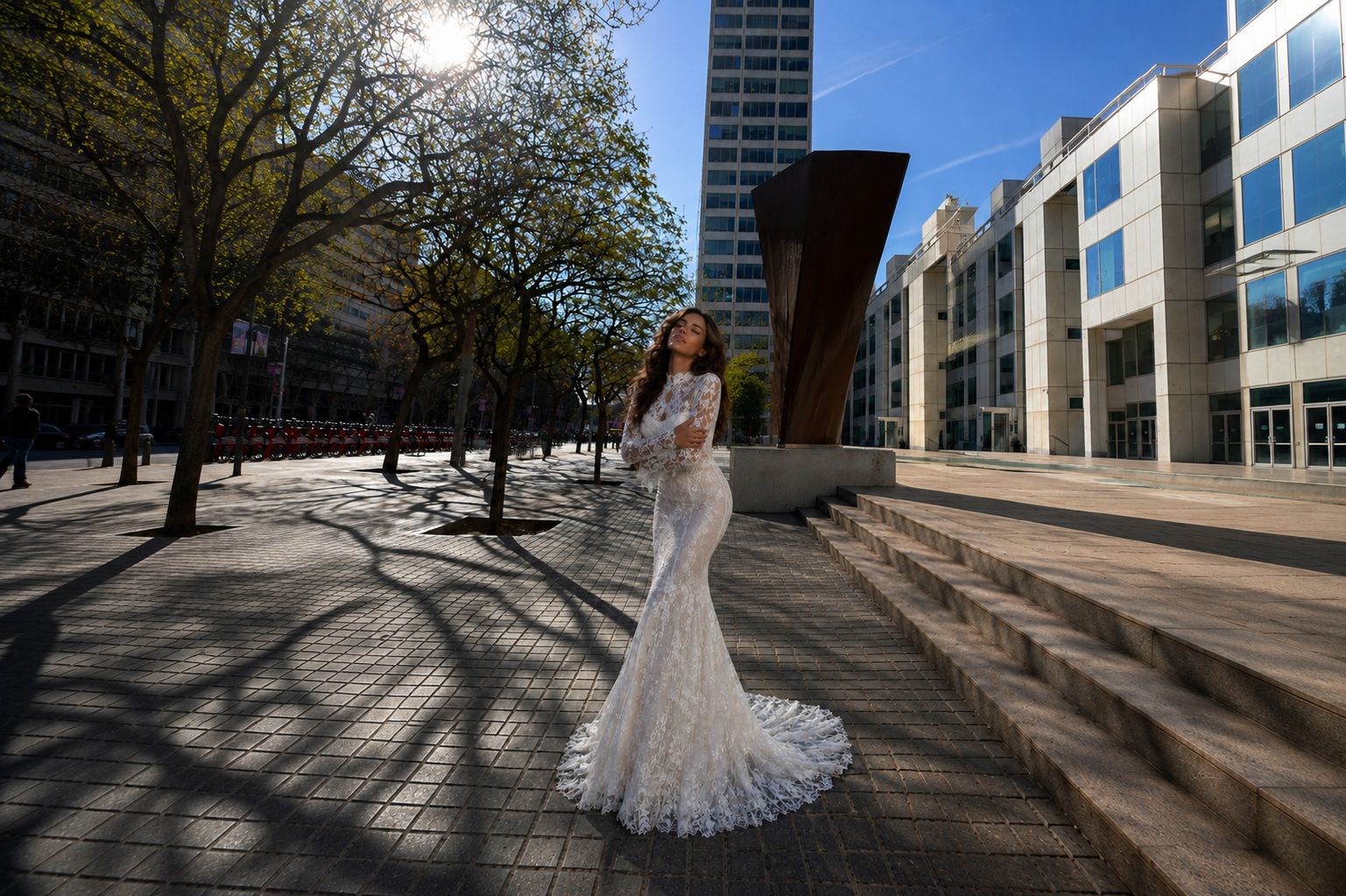 Donna con abito da sposa bianco aderente in pizzo in un'area urbana soleggiata con alberi e edifici.