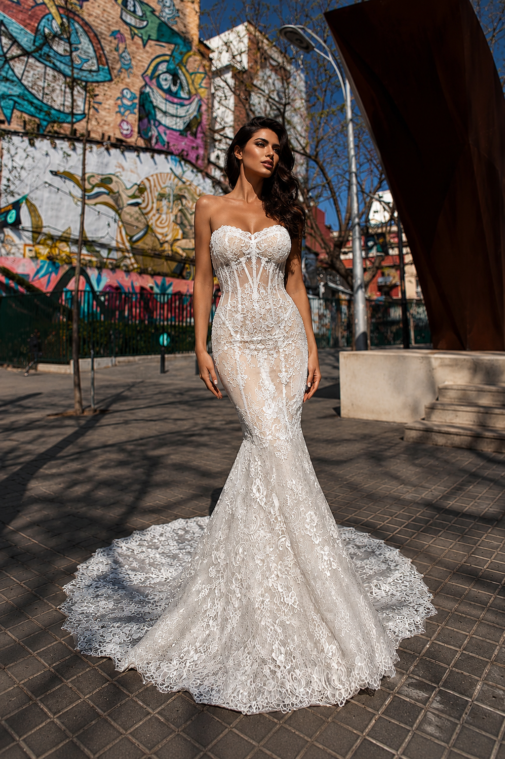 Donna in abito da sposa bianco con pizzo dettagliato in città con murales colorati sullo sfondo.