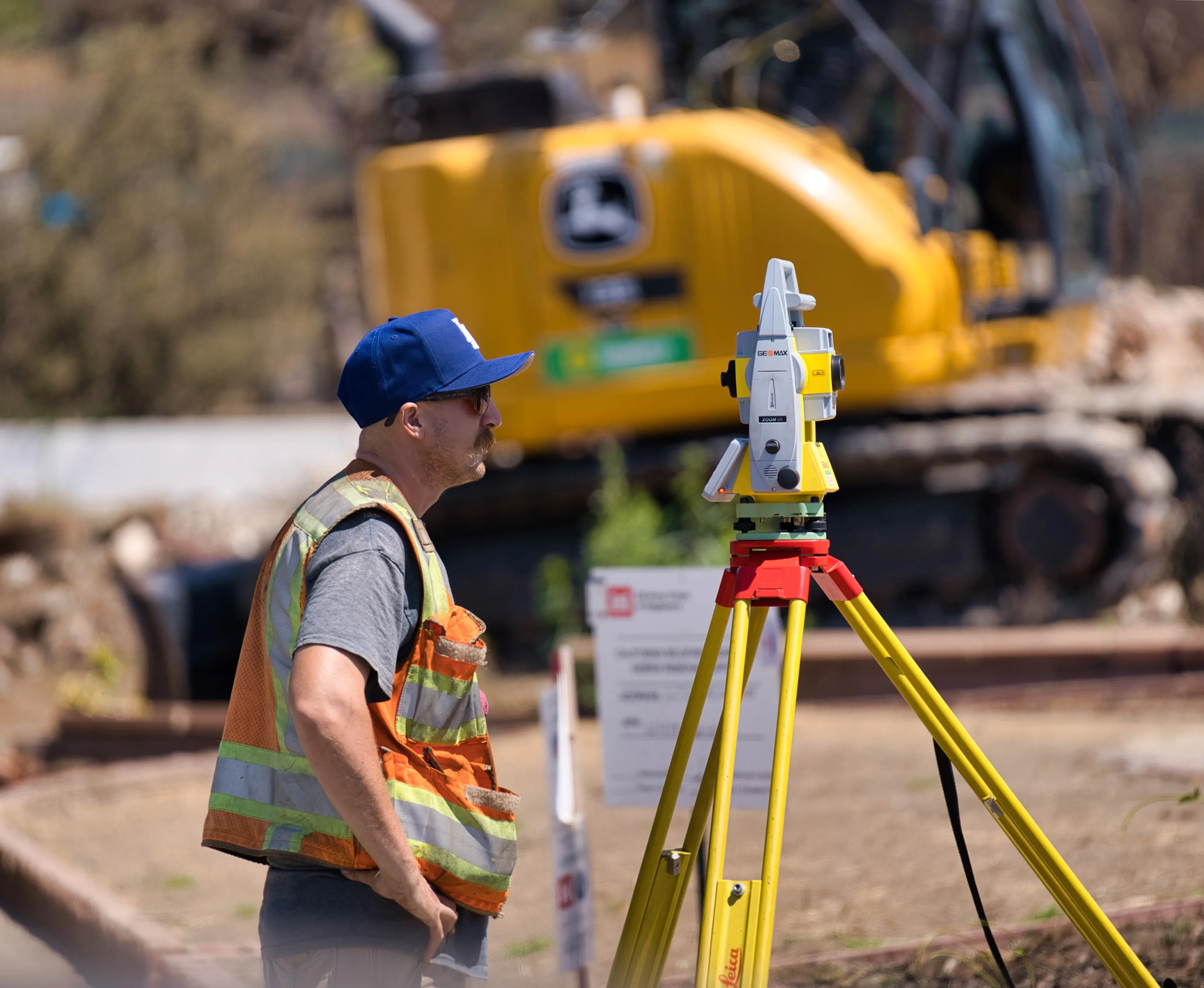 Builoff Surveyor in safety vest and cap operating a surveying instrument on a tripod at a construction site in Altadena with excavator in the background.