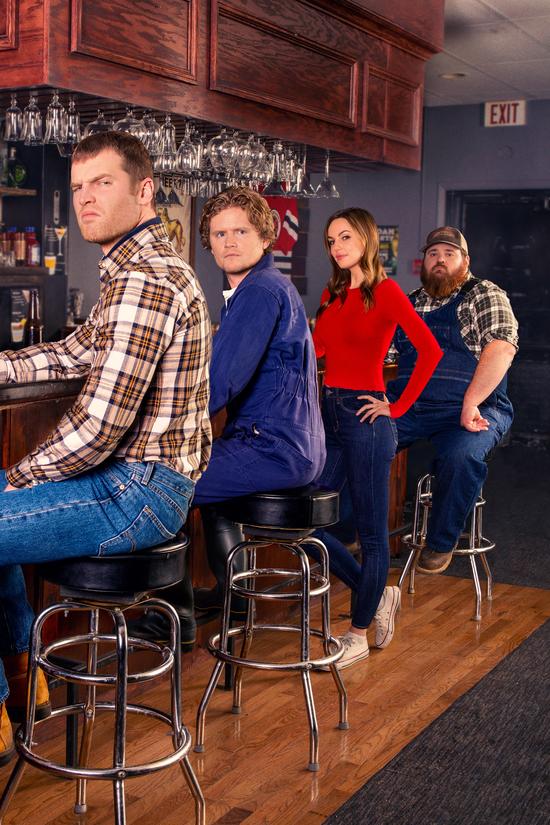 Four people sitting and standing at a bar with wooden stools and glasses hanging overhead, including a man in a plaid shirt, a man in blue coveralls, a woman in a red top, and a man in overalls and a cap.